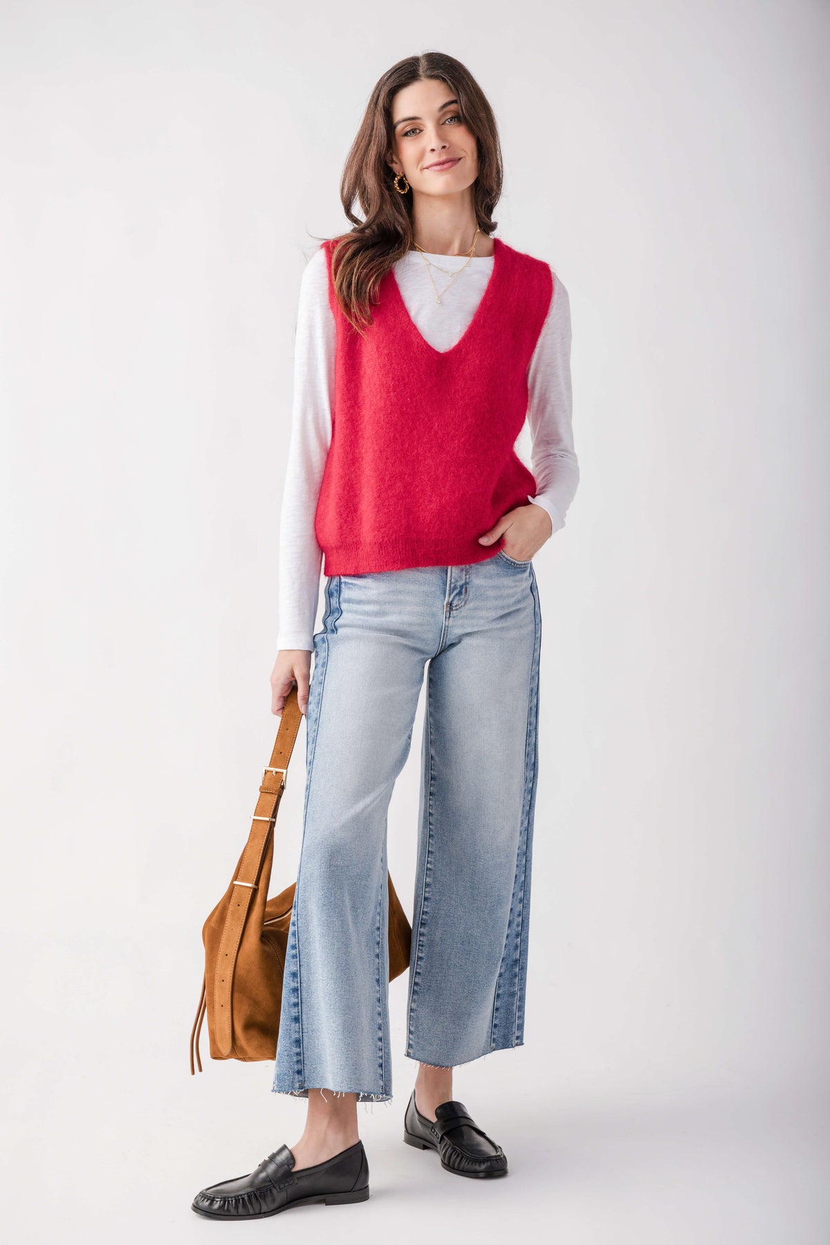 A woman modeling a bright red knit sweater vest over a white long-sleeve shirt, paired with light-wash wide-leg jeans, black loafers, and carrying a tan shoulder bag against a light gray studio background.