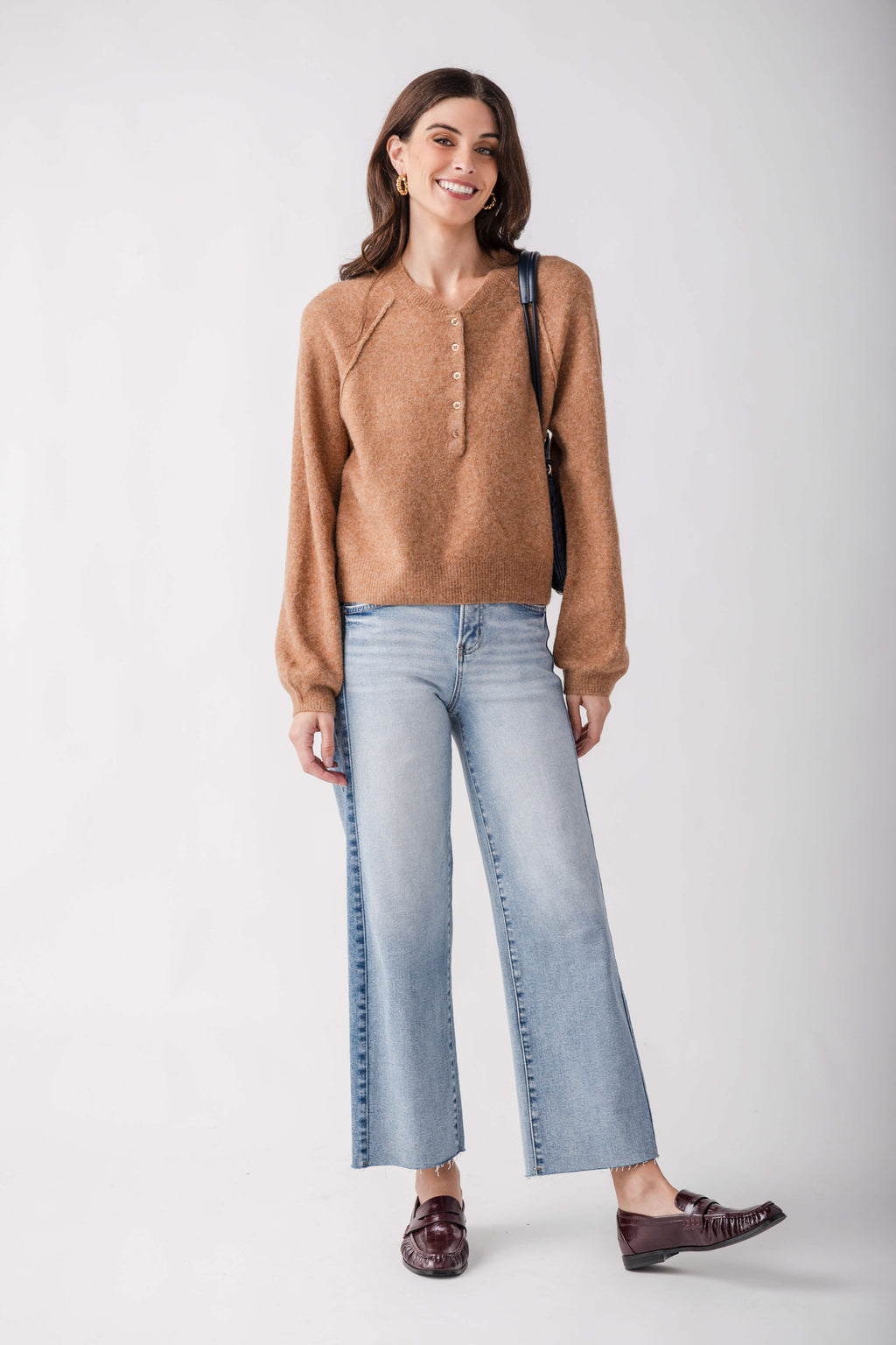 A woman modeling a light brown knit sweater, light-wash wide-leg cropped jeans, and dark brown loafers against a plain light gray studio background.