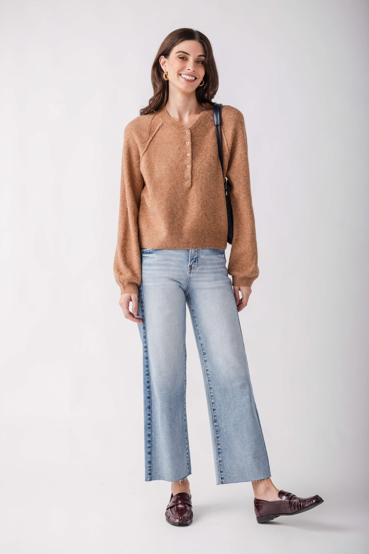 A woman modeling a light brown knit sweater, light-wash wide-leg cropped jeans, and dark brown loafers against a plain light gray studio background.
