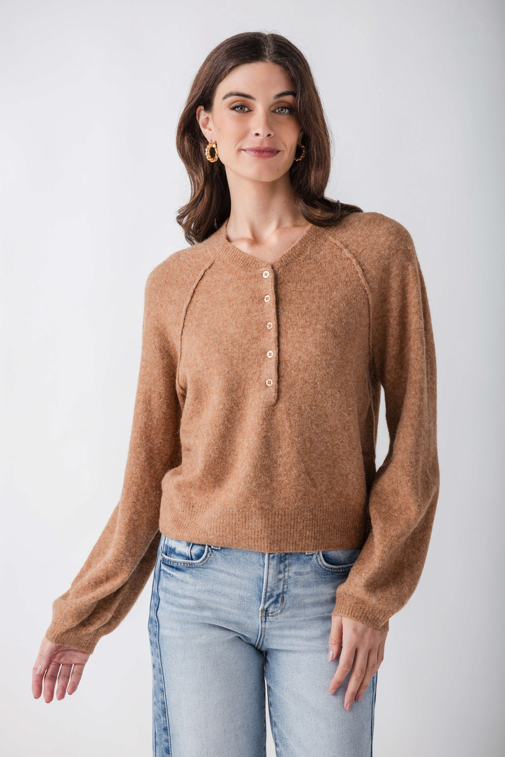 Tan henley sweater with button front placket and long sleeves worn with light blue jeans.
