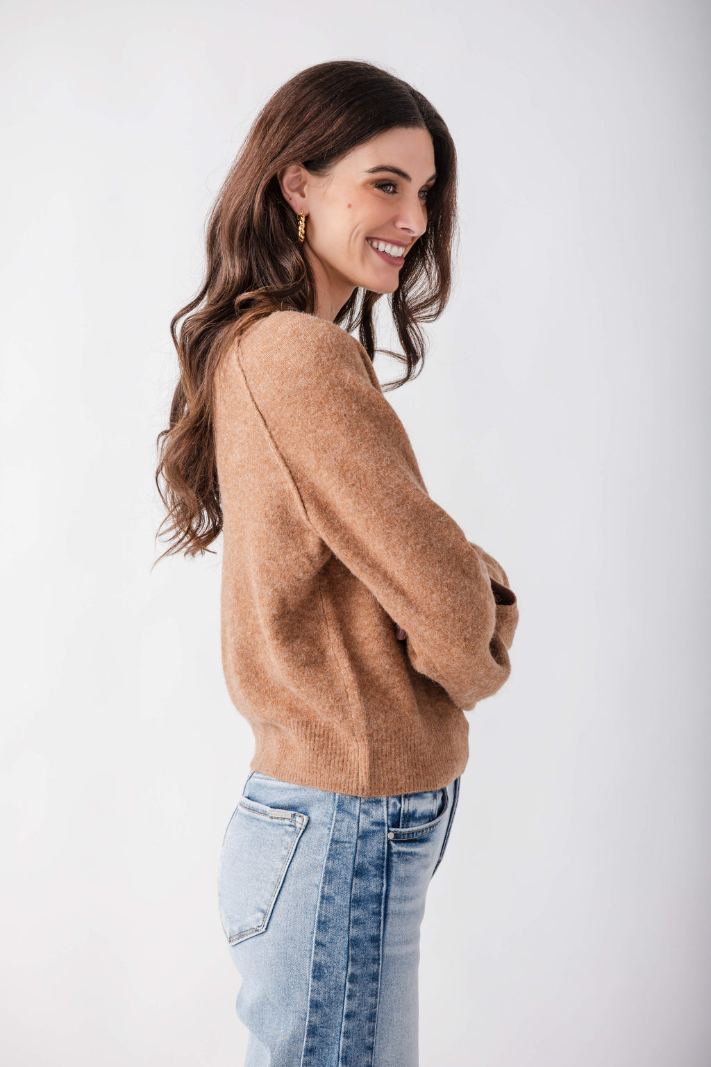 Woman wearing a tan henley sweater with long sleeves paired with light blue jeans, shown from the side.
