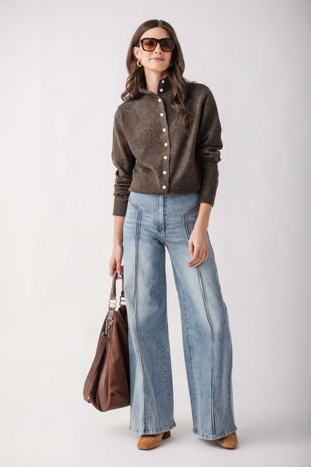A woman modeling a brown button-front knit cardigan, light-wash wide-leg denim jeans, and brown shoes, holding a brown leather handbag against a plain light gray studio background.