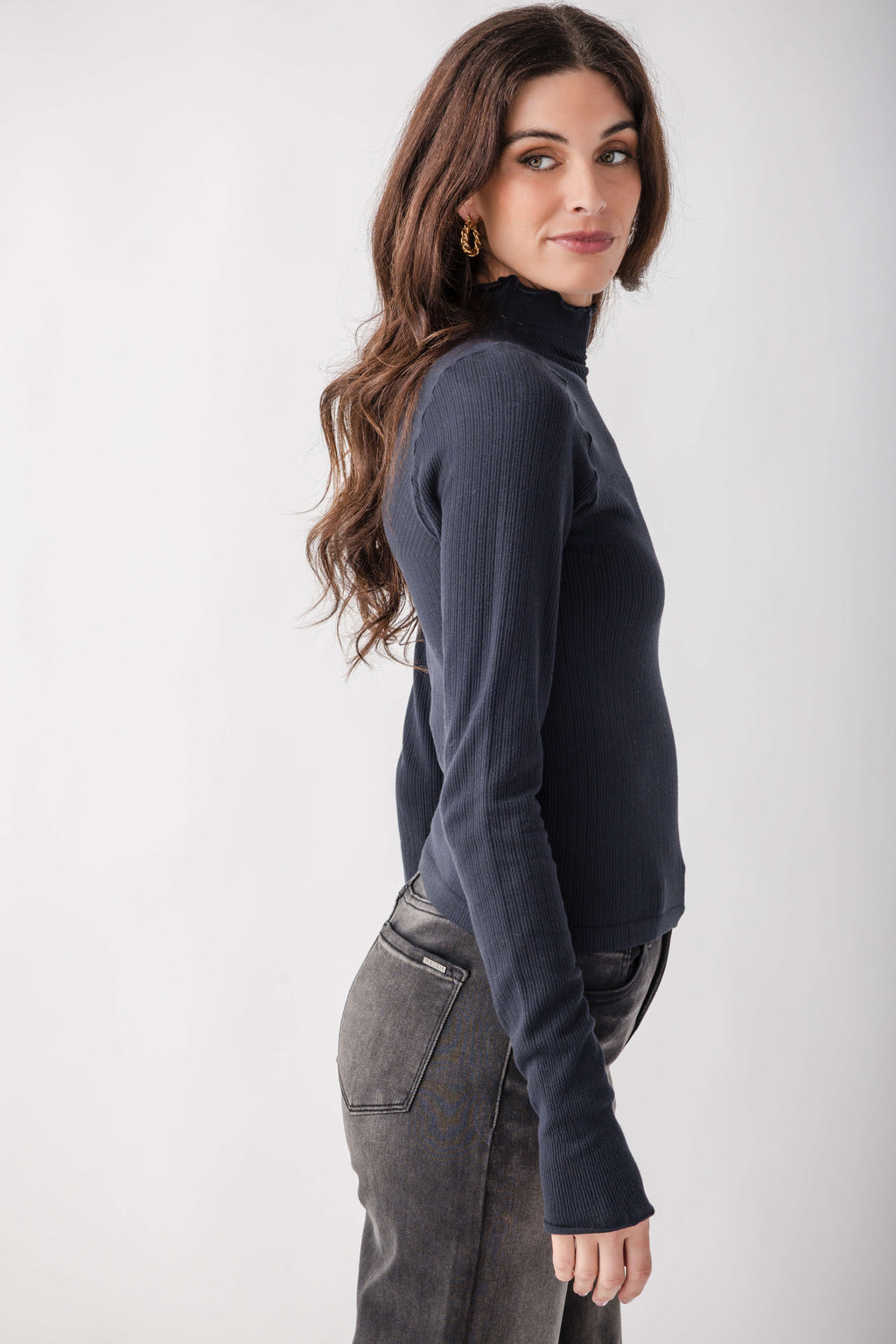 A woman modeling a dark navy ribbed turtleneck sweater, paired with gray jeans, against a plain light gray studio background.