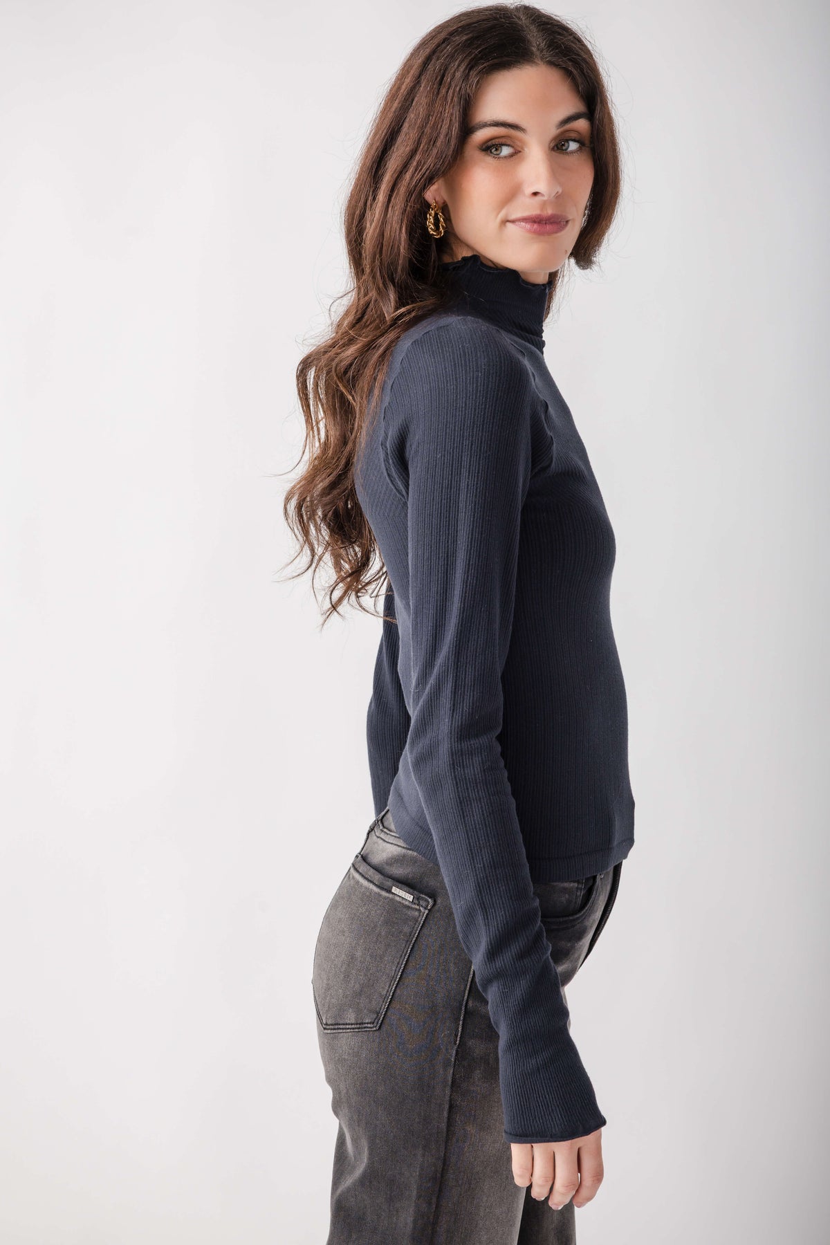 A woman modeling a dark navy ribbed turtleneck sweater, paired with gray jeans, against a plain light gray studio background.