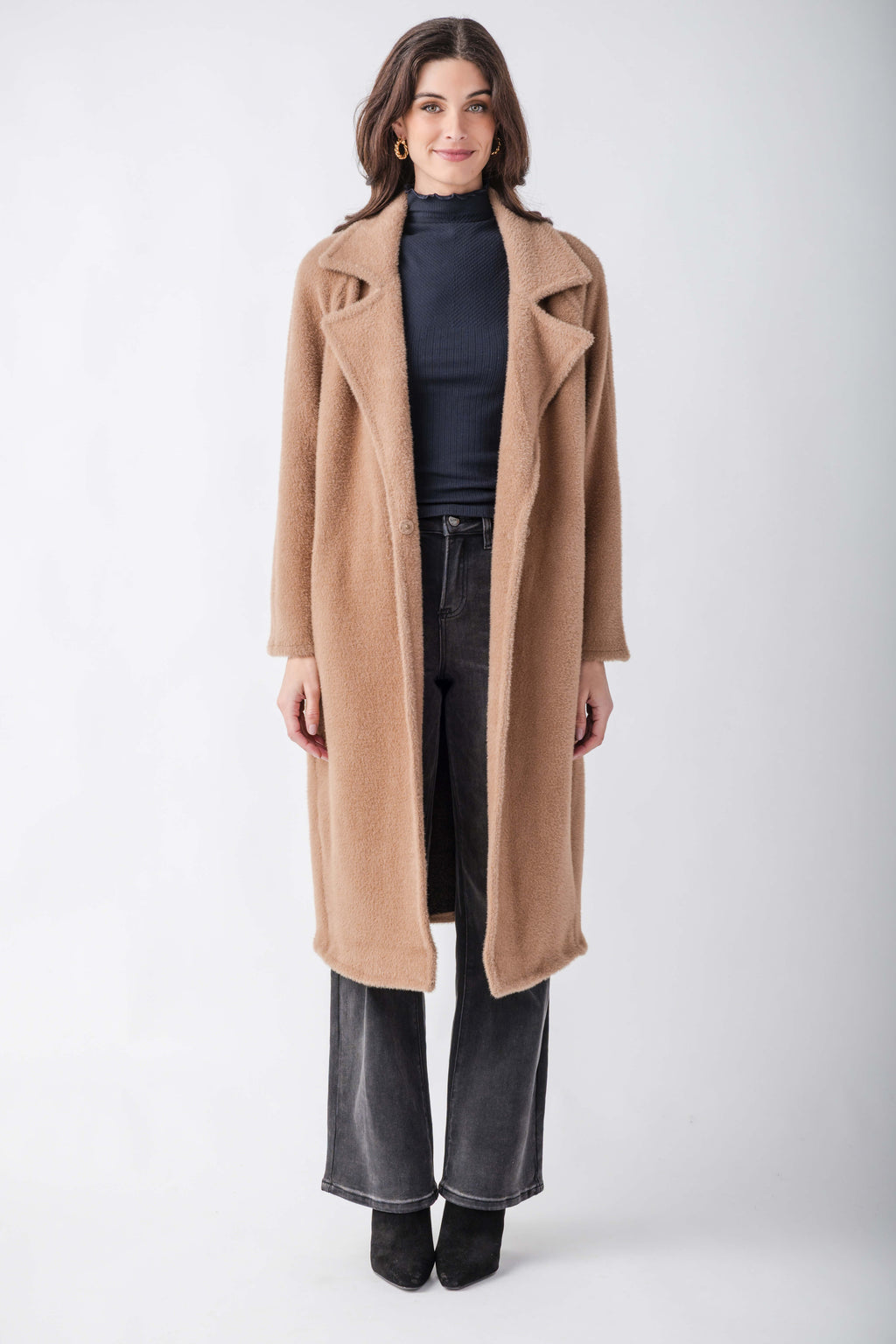 A woman modeling a long camel-colored wool coat over a dark navy turtleneck, black wide-leg jeans, and black ankle boots, photographed against a neutral light gray studio background.