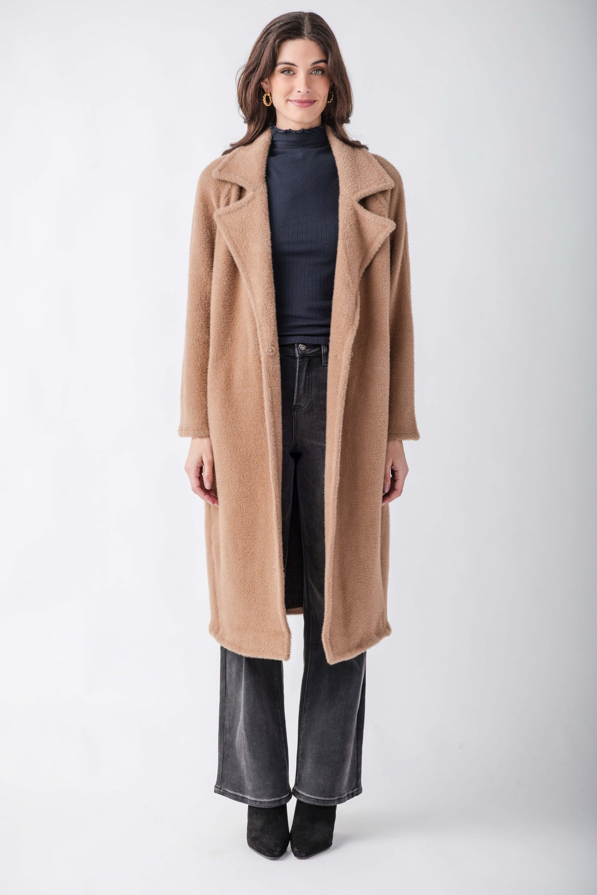 A woman modeling a long camel-colored wool coat over a dark navy turtleneck, black wide-leg jeans, and black ankle boots, photographed against a neutral light gray studio background.