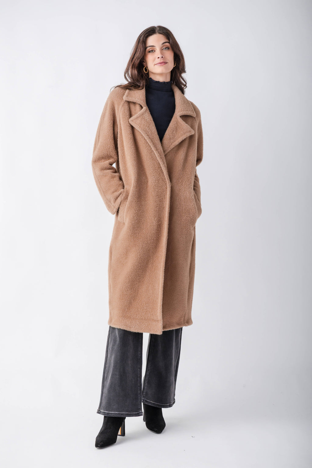 A woman modeling a long camel-colored wool coat over a black turtleneck, wide-leg charcoal gray pants, and black ankle boots, photographed against a seamless light gray studio background.
