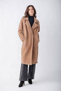 A woman modeling a long camel-colored wool coat over a black turtleneck, wide-leg charcoal gray pants, and black ankle boots, photographed against a seamless light gray studio background.
