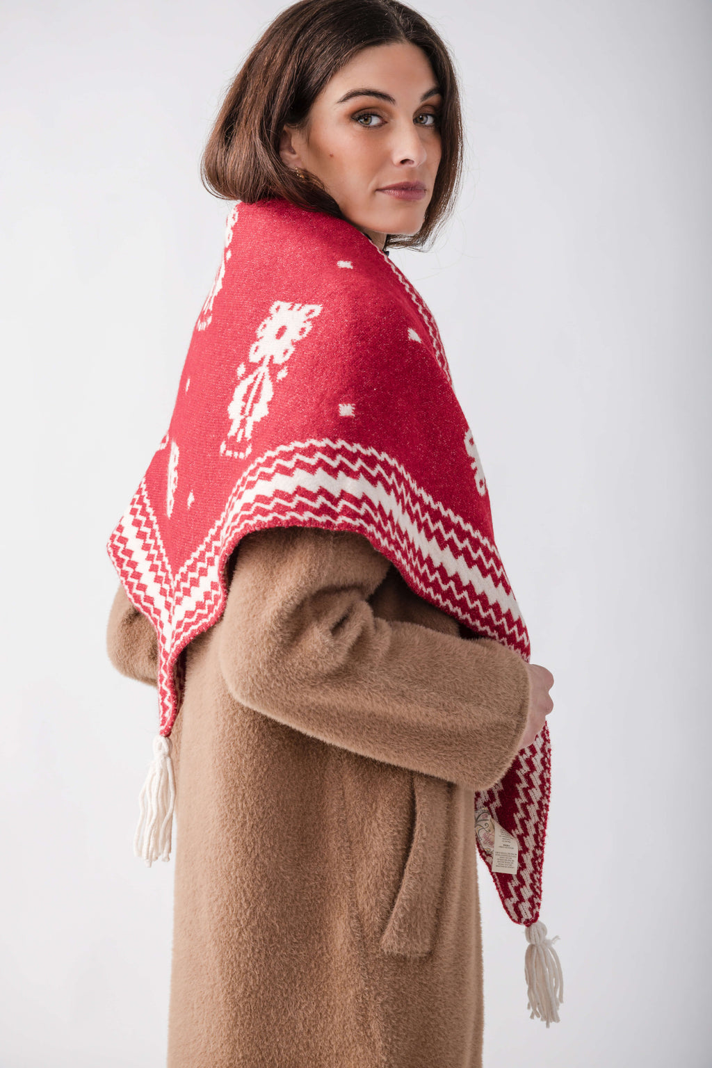 Woman wearing red and white patterned shawl over beige coat with white fringe trim.