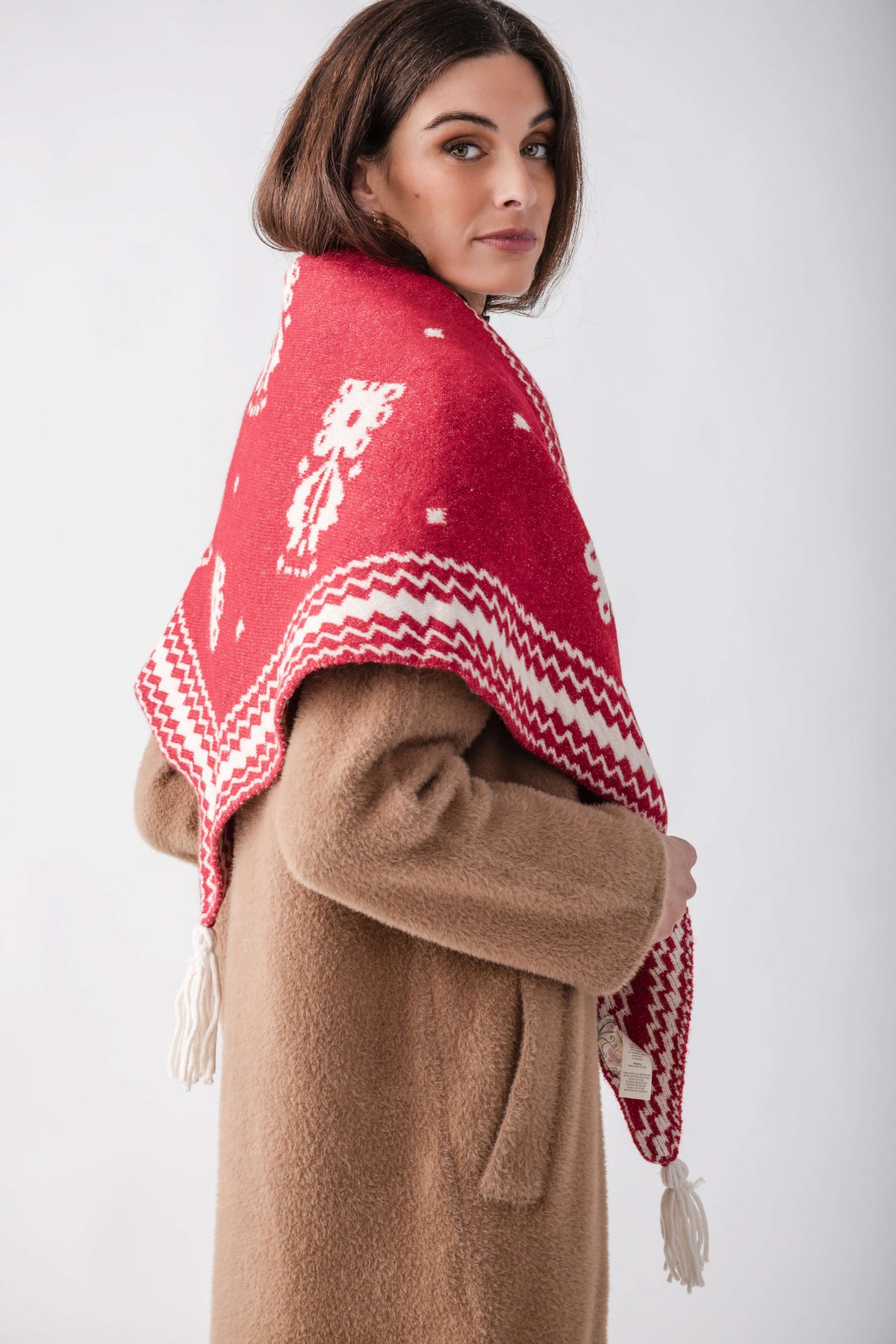 Woman wearing red and white patterned shawl over beige coat with white fringe trim.