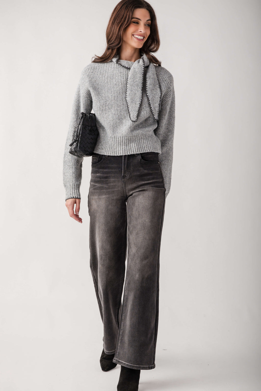 A woman modeling a light gray ribbed knit sweater with a decorative tie detail at the collar, paired with dark gray wide-leg jeans and black ankle boots, against a plain light gray studio background.