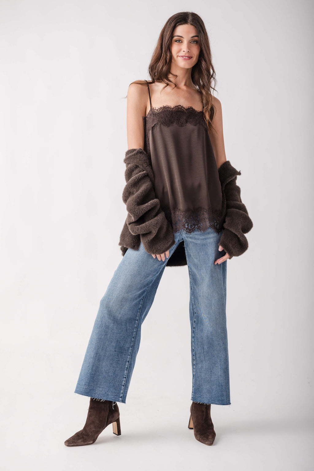 A model wearing a dark brown satin camisole top with lace trim, a matching dark brown fuzzy cropped cardigan, wide-leg blue jeans, and brown suede boots, posed against a light gray studio background.