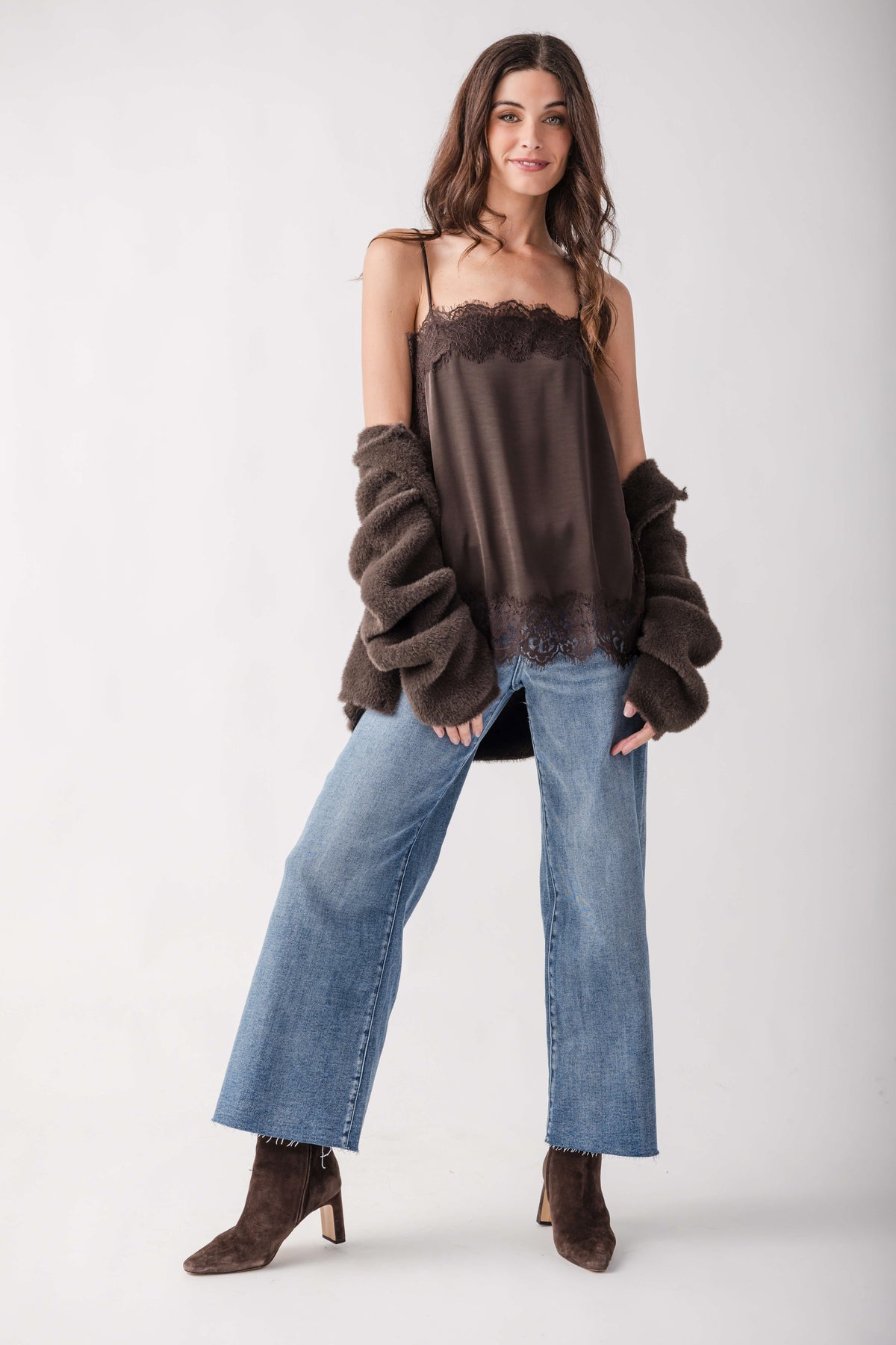 A model wearing a dark brown satin camisole top with lace trim, a matching dark brown fuzzy cropped cardigan, wide-leg blue jeans, and brown suede boots, posed against a light gray studio background.