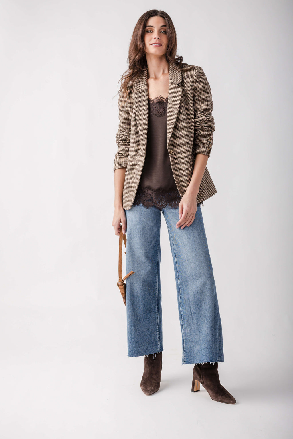 A woman models a brown textured blazer over a dark brown lace-trimmed top, paired with wide-leg blue jeans and brown suede ankle boots, holding a small brown handbag against a light gray studio background.