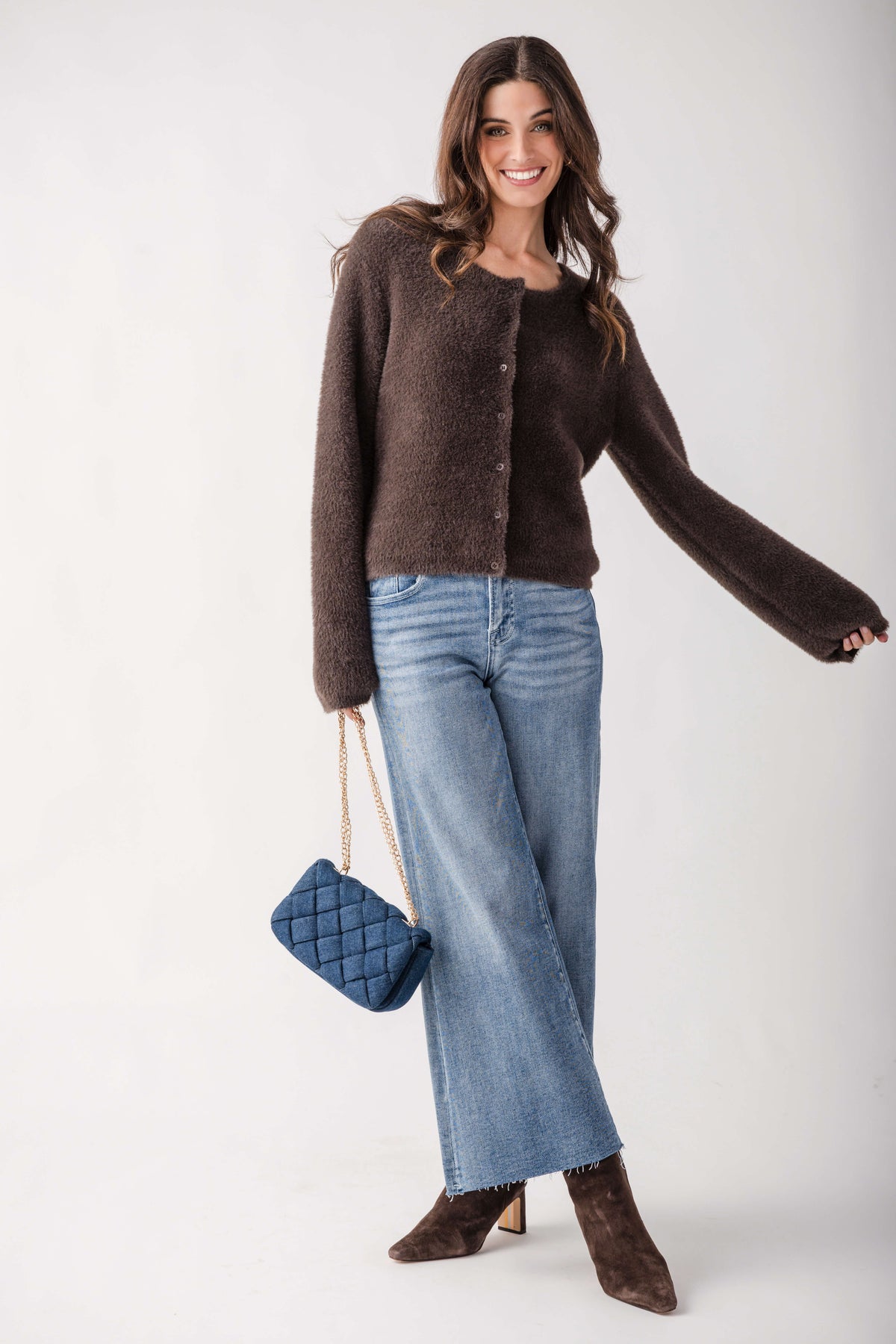 A woman modeling a dark brown fuzzy knit cardigan, blue wide-leg jeans, brown suede ankle boots, and a blue quilted handbag against a plain white studio background.