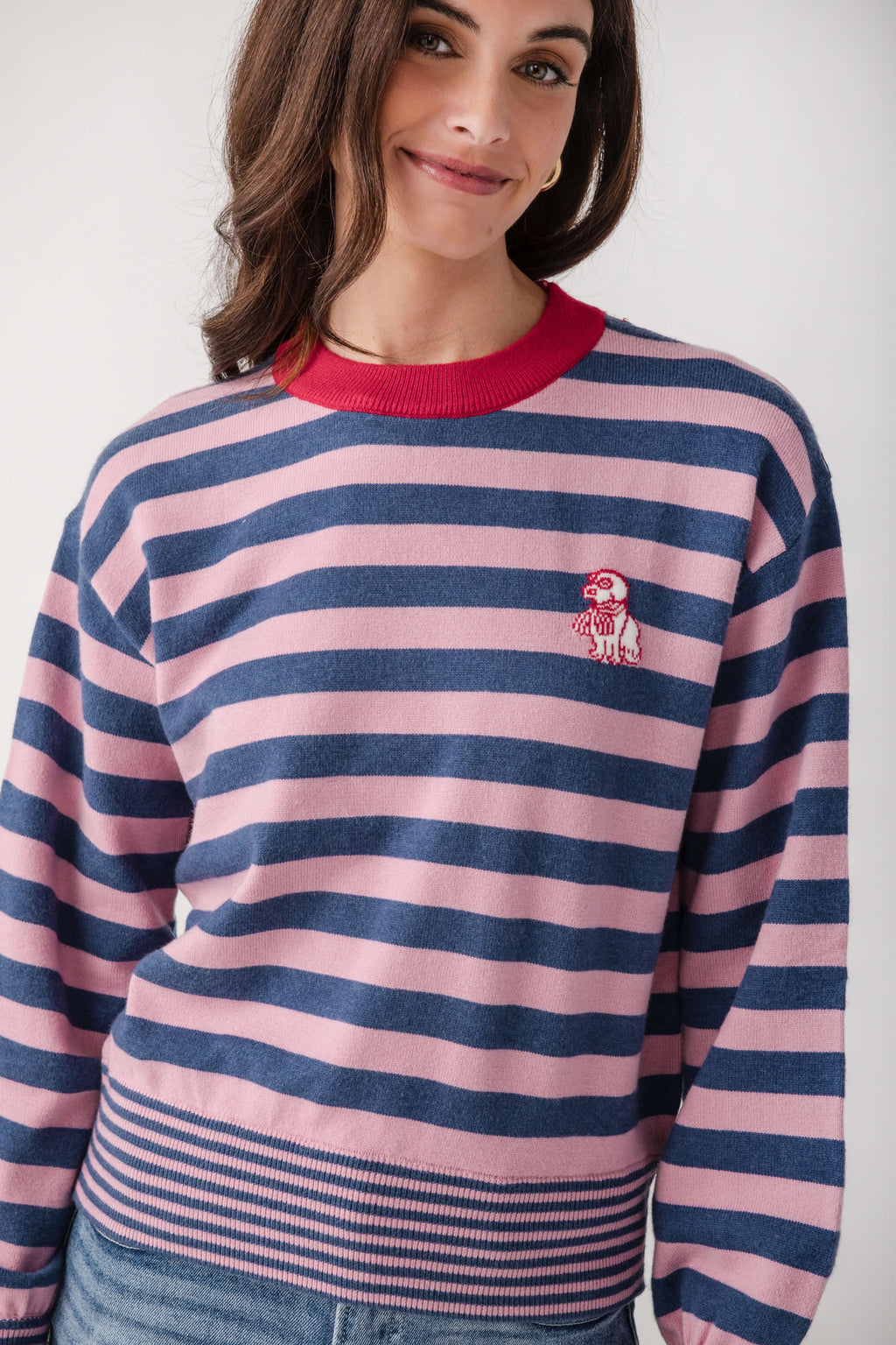 A woman wearing a pink and navy blue horizontally striped crewneck sweater with a red collar and a small embroidered dog logo on the chest, paired with blue jeans against a light gray background.