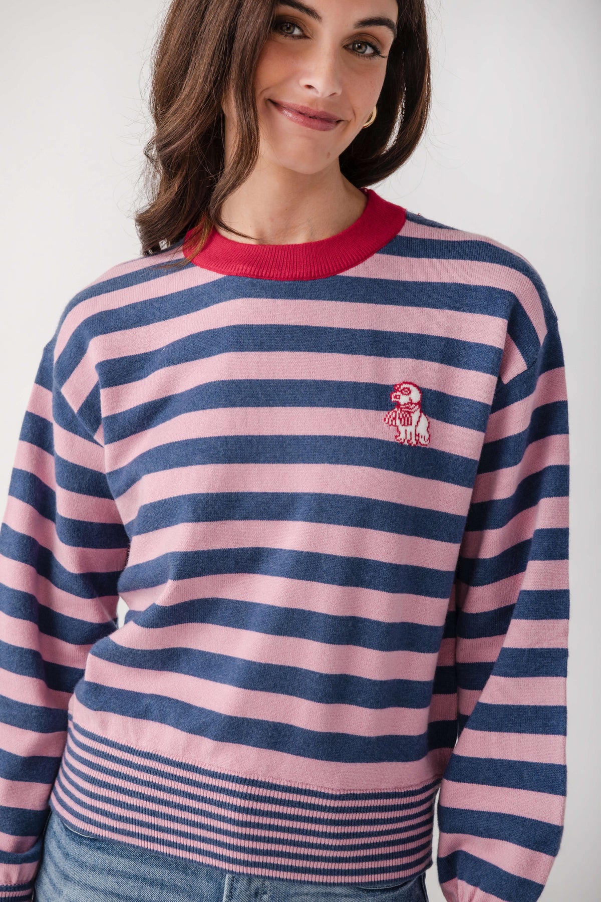 A woman wearing a pink and navy blue horizontally striped crewneck sweater with a red collar and a small embroidered dog logo on the chest, paired with blue jeans against a light gray background.