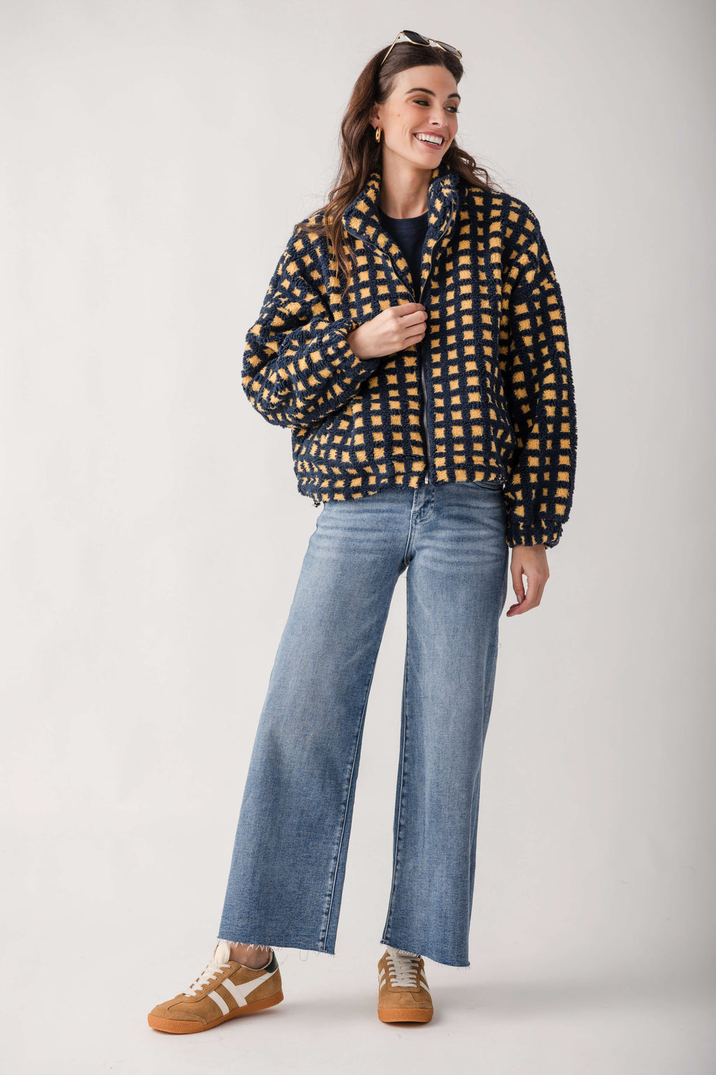 A woman modeling a navy blue and mustard yellow checkered puffer jacket, paired with wide-leg blue jeans and brown sneakers, against a plain light gray studio background.