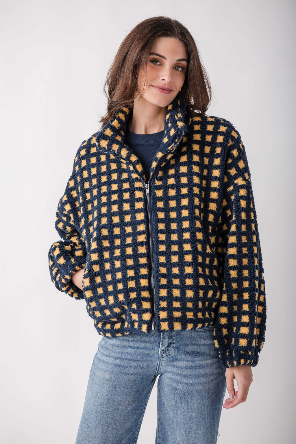 Woman wearing a navy and gold checkered sherpa fleece jacket with a full-length zipper over a dark shirt and light blue jeans