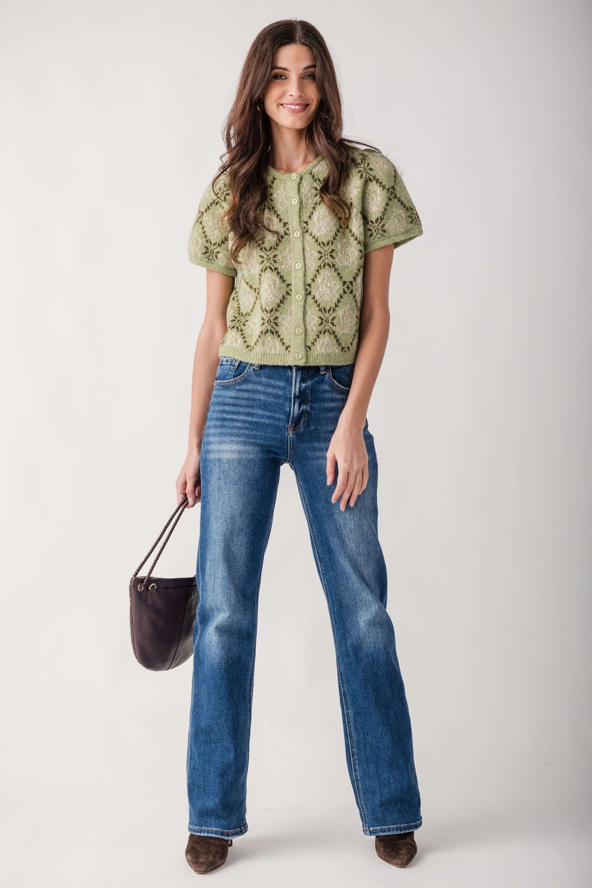 A woman modeling a light olive green short-sleeve knit top with a textured geometric pattern, paired with blue jeans and holding a dark purple handbag against a light gray studio background.