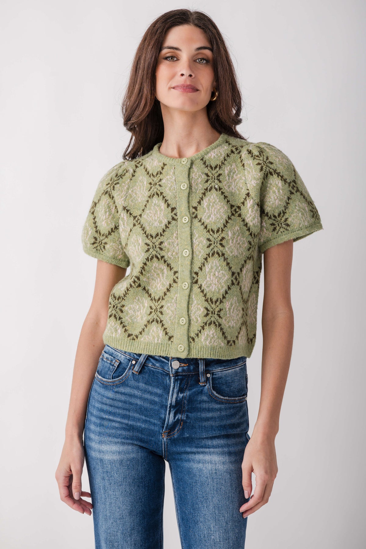 A woman modeling a short-sleeve knit cardigan with a green and beige geometric diamond pattern, paired with blue jeans, against a light gray studio background.