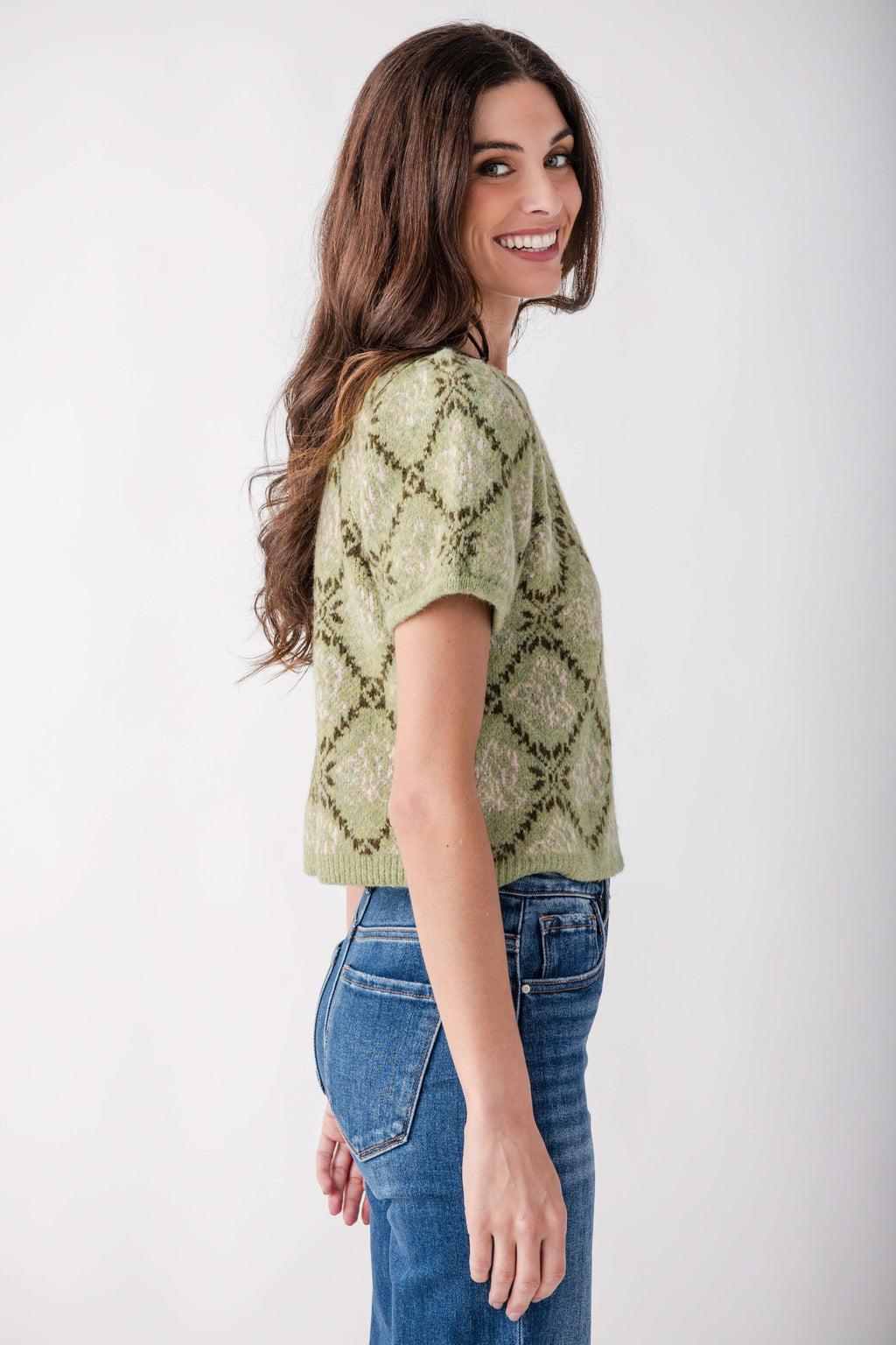 A woman wearing a light olive green short-sleeve knit top with a dark green geometric diamond pattern, paired with blue jeans, photographed against a plain white studio background.