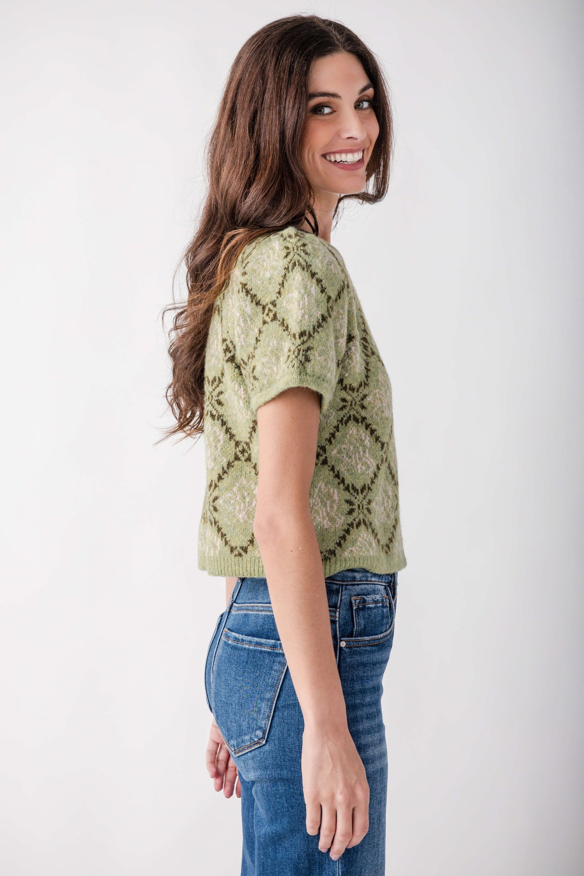 A woman wearing a light olive green short-sleeve knit top with a dark green geometric diamond pattern, paired with blue jeans, photographed against a plain white studio background.