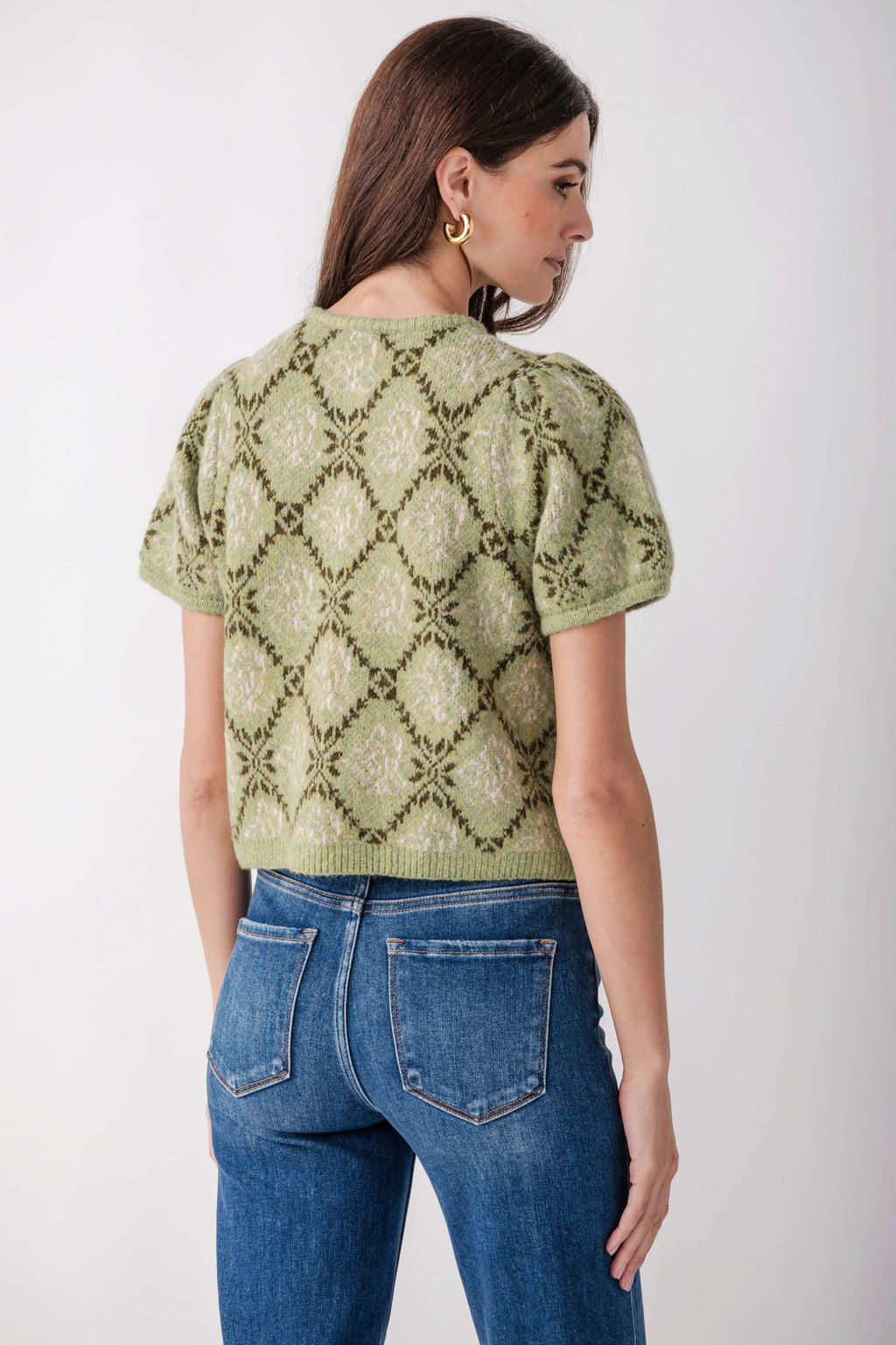 A woman shown from the back wearing a light olive green short-sleeve knit top with a darker olive green geometric diamond pattern, paired with blue jeans, against a plain light gray background.