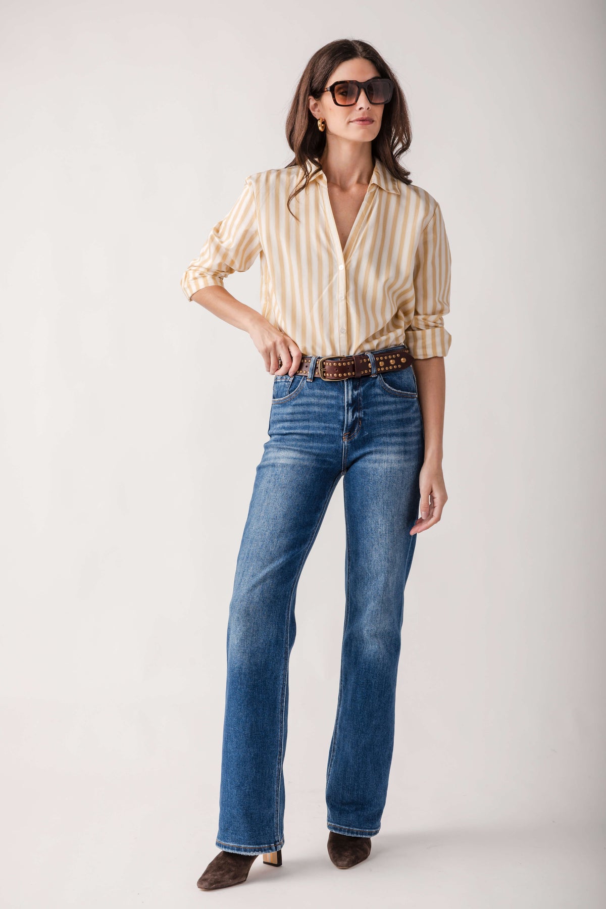 A woman modeling a cream and tan vertically striped button-down shirt, blue jeans, a brown belt, and brown shoes against a light gray studio background.