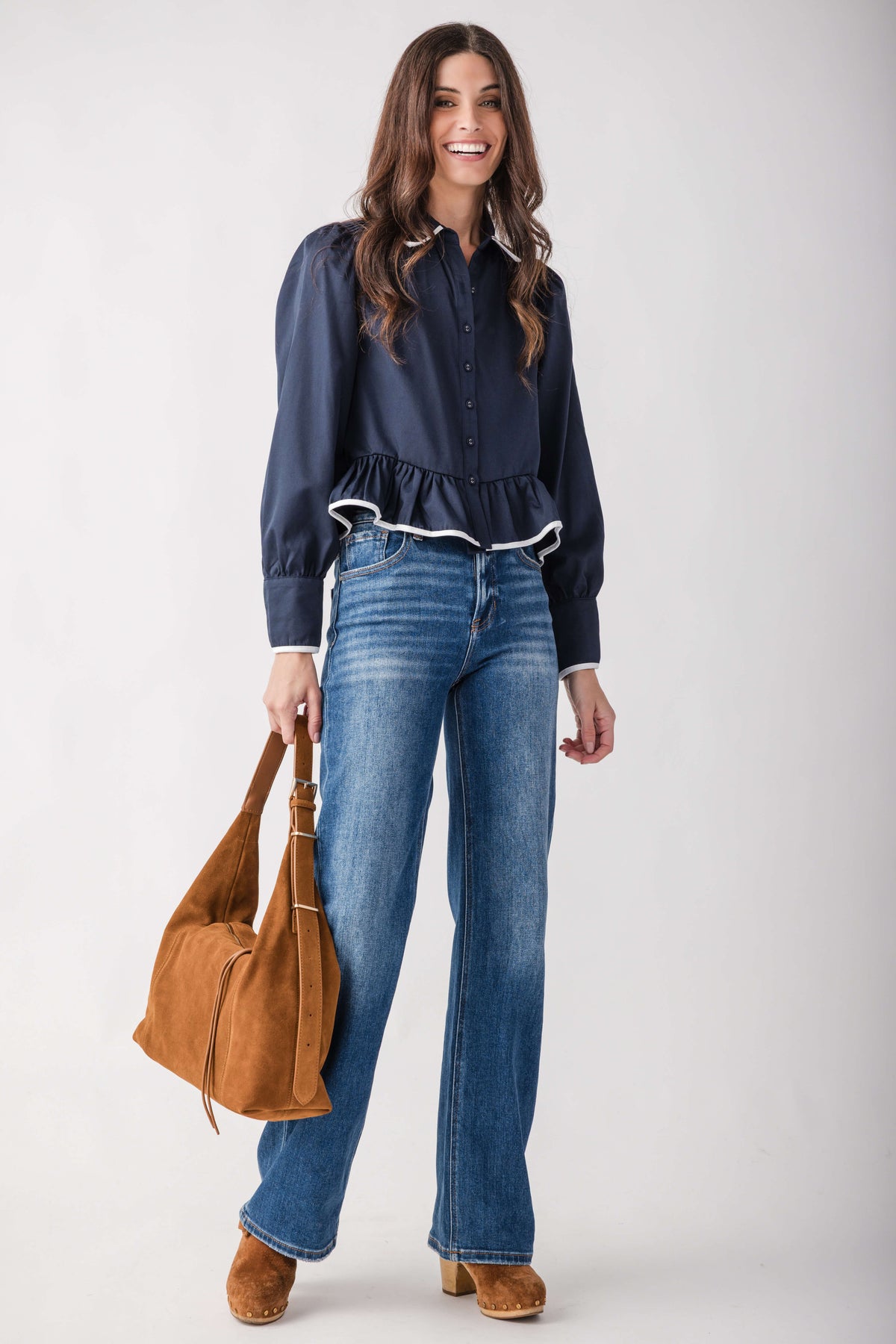 A woman modeling a navy blue long-sleeve blouse with white piping, blue jeans, and brown ankle boots, holding a brown suede shoulder bag against a light gray studio background.