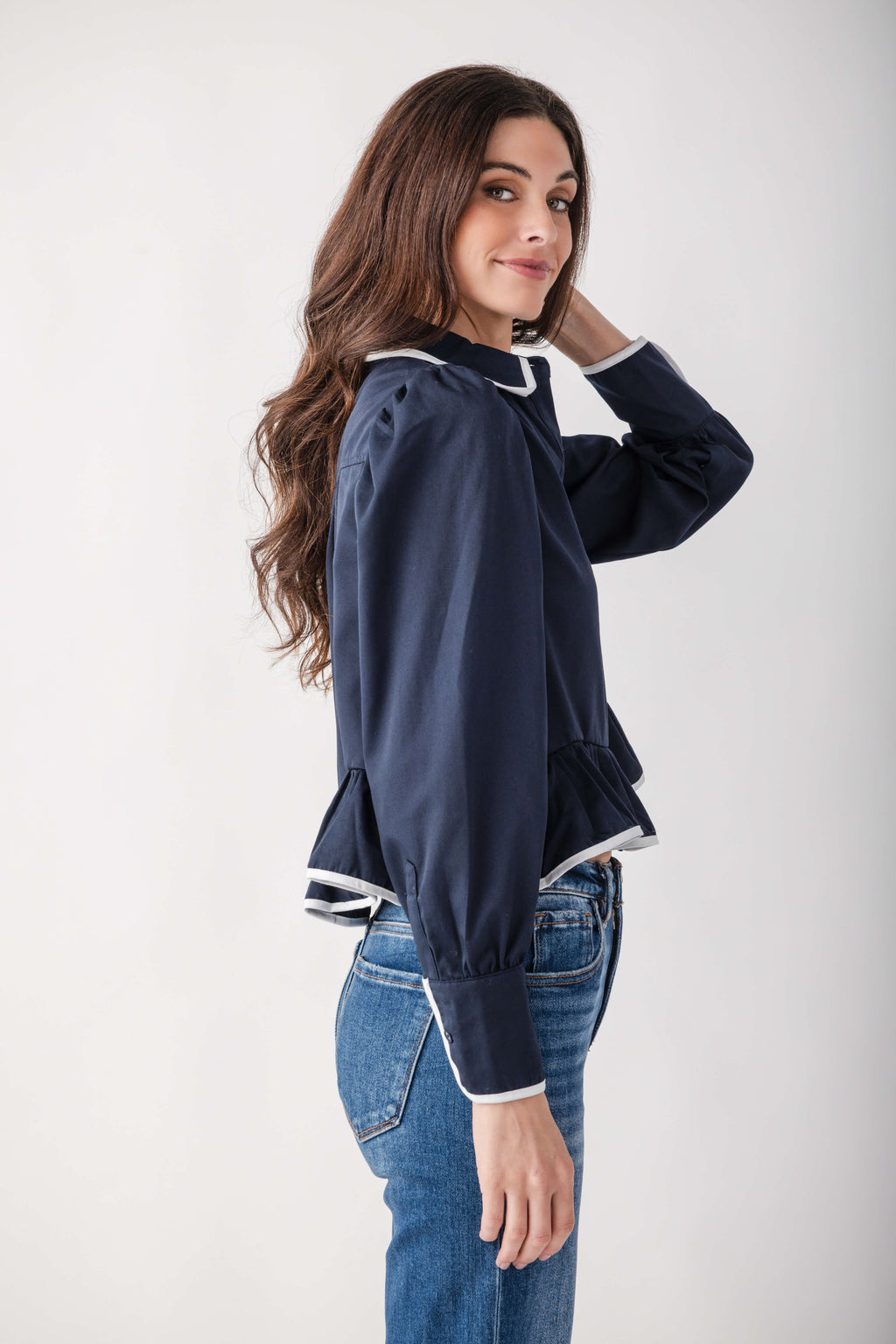 A woman wearing a navy blue long-sleeve blouse with white contrast trim and blue jeans, photographed against a plain light gray studio background.