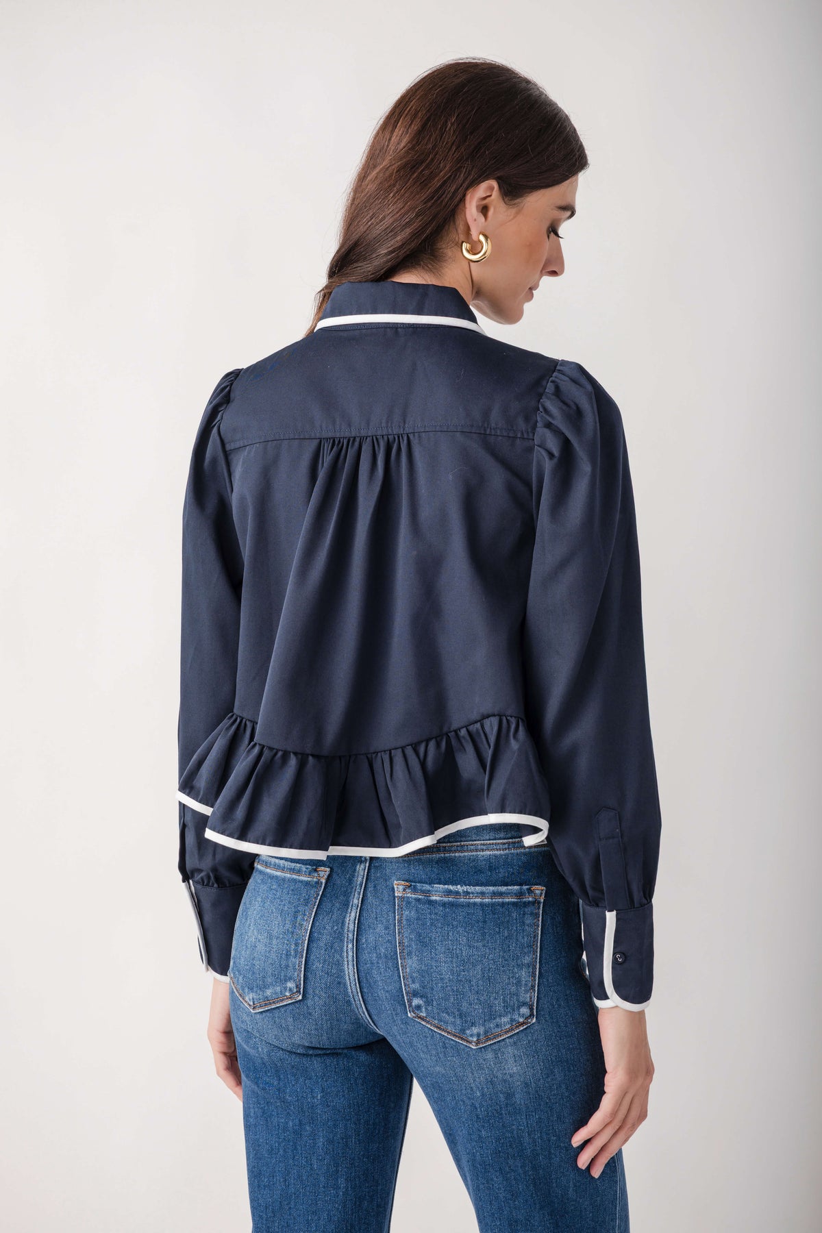 A woman shown from the back wearing a navy blue long-sleeve blouse with white piping trim and blue jeans, photographed against a light gray studio background.