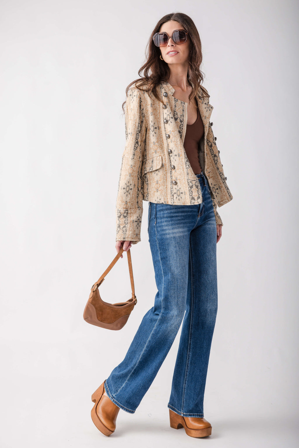 A woman modeling a beige patterned jacket, blue wide-leg jeans, a brown top, sunglasses, and brown clogs, holding a brown handbag against a light gray studio background.