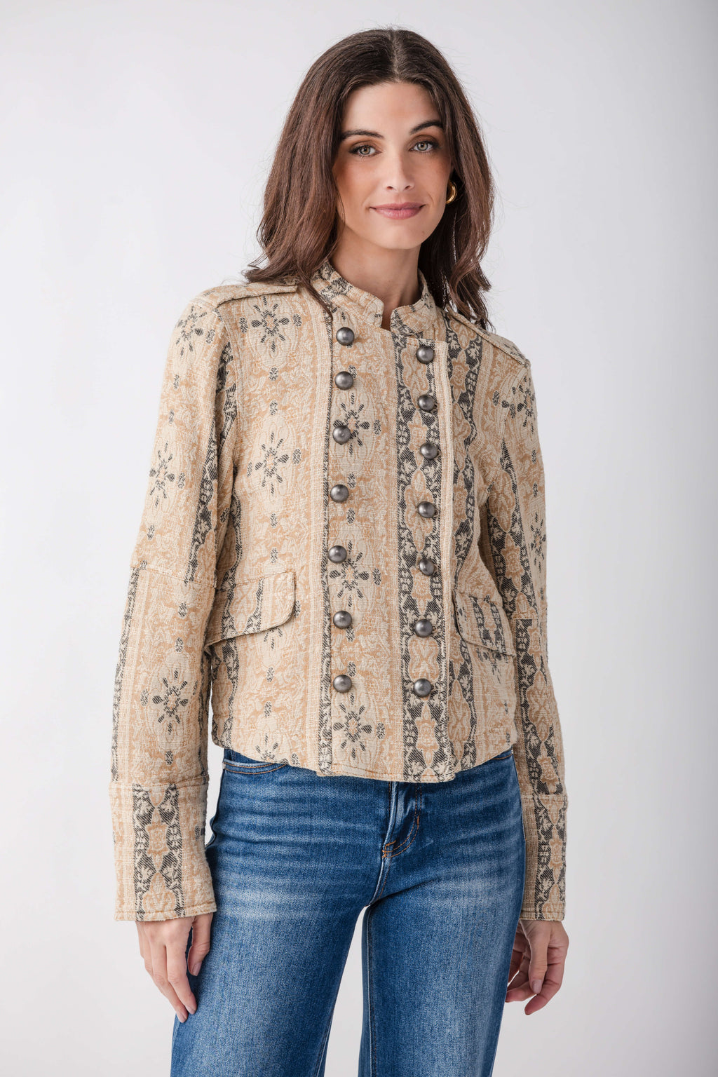 A woman modeling a cropped, long-sleeve jacket with a beige and brown floral pattern, paired with blue jeans, against a light gray studio background.