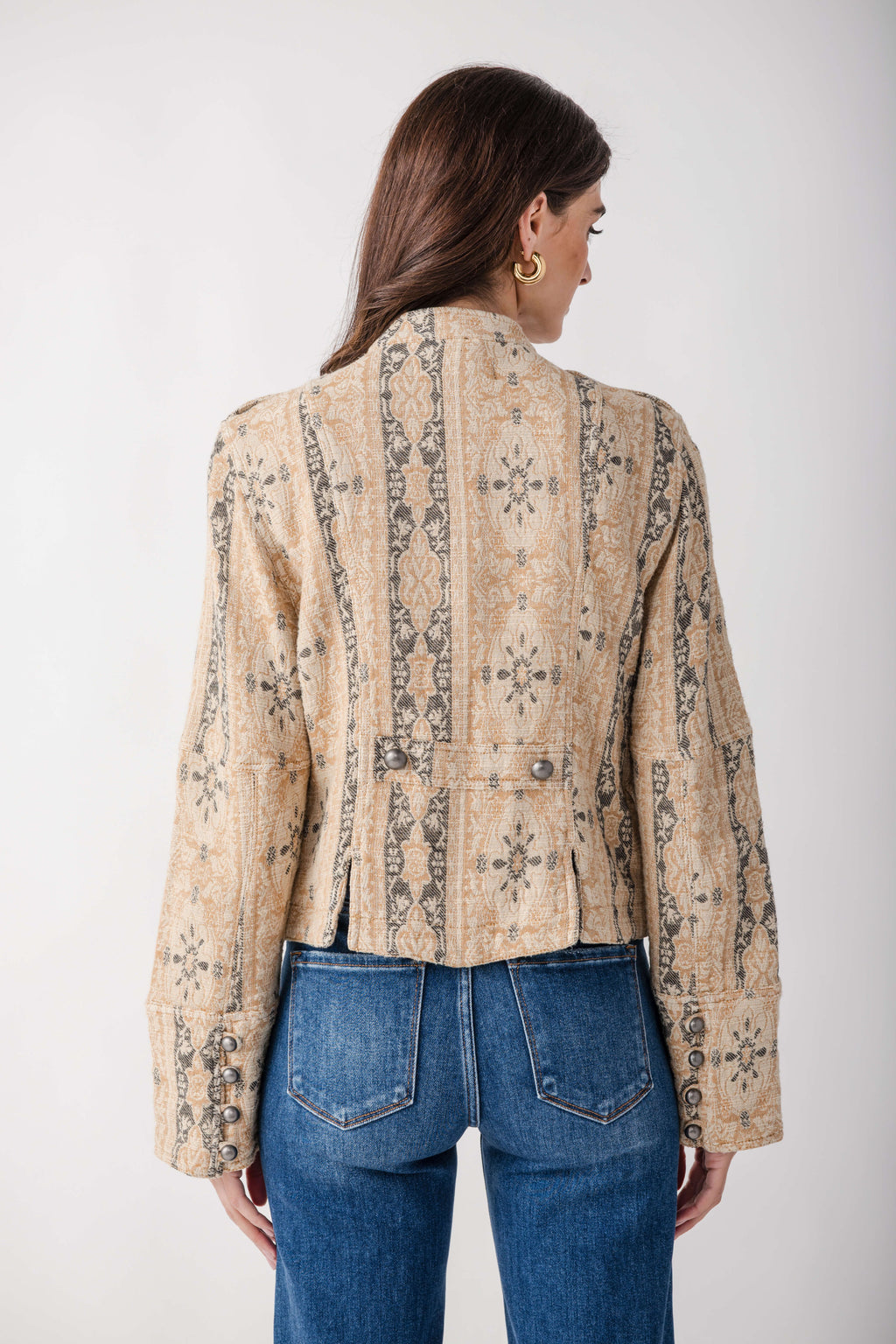 A woman viewed from the back wearing a cropped beige jacket with a dark brown geometric and tribal-style pattern, paired with blue jeans, against a plain light gray background.
