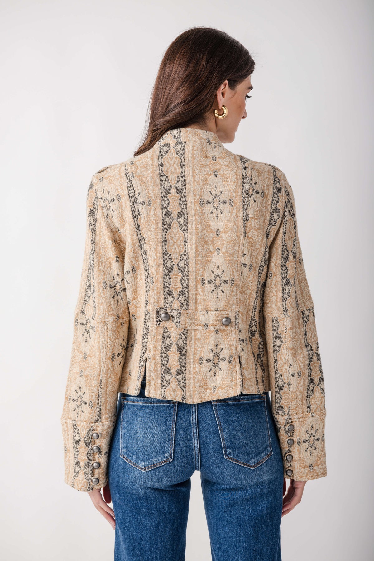 A woman viewed from the back wearing a cropped beige jacket with a dark brown geometric and tribal-style pattern, paired with blue jeans, against a plain light gray background.
