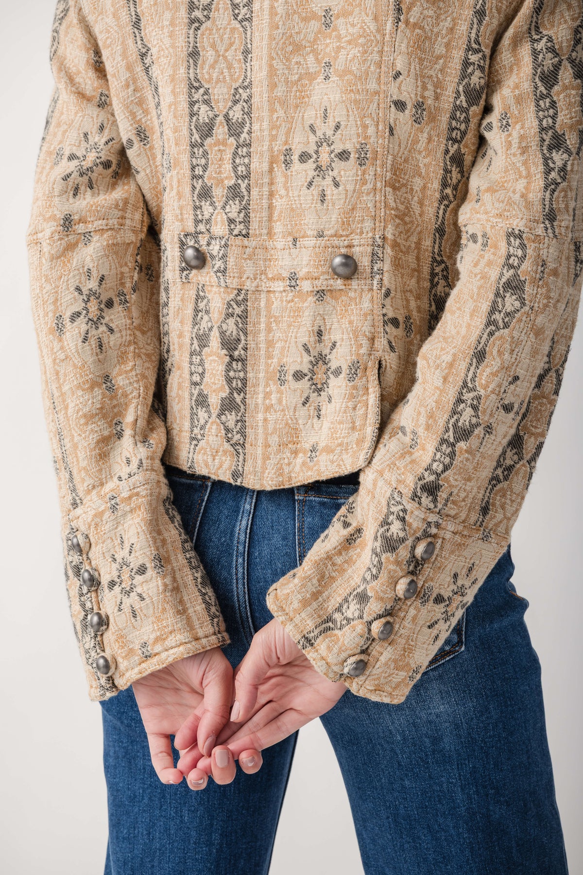 A close-up of a person wearing a cropped, long-sleeve quilted jacket with a beige and brown geometric pattern, paired with blue jeans, against a plain light background.
