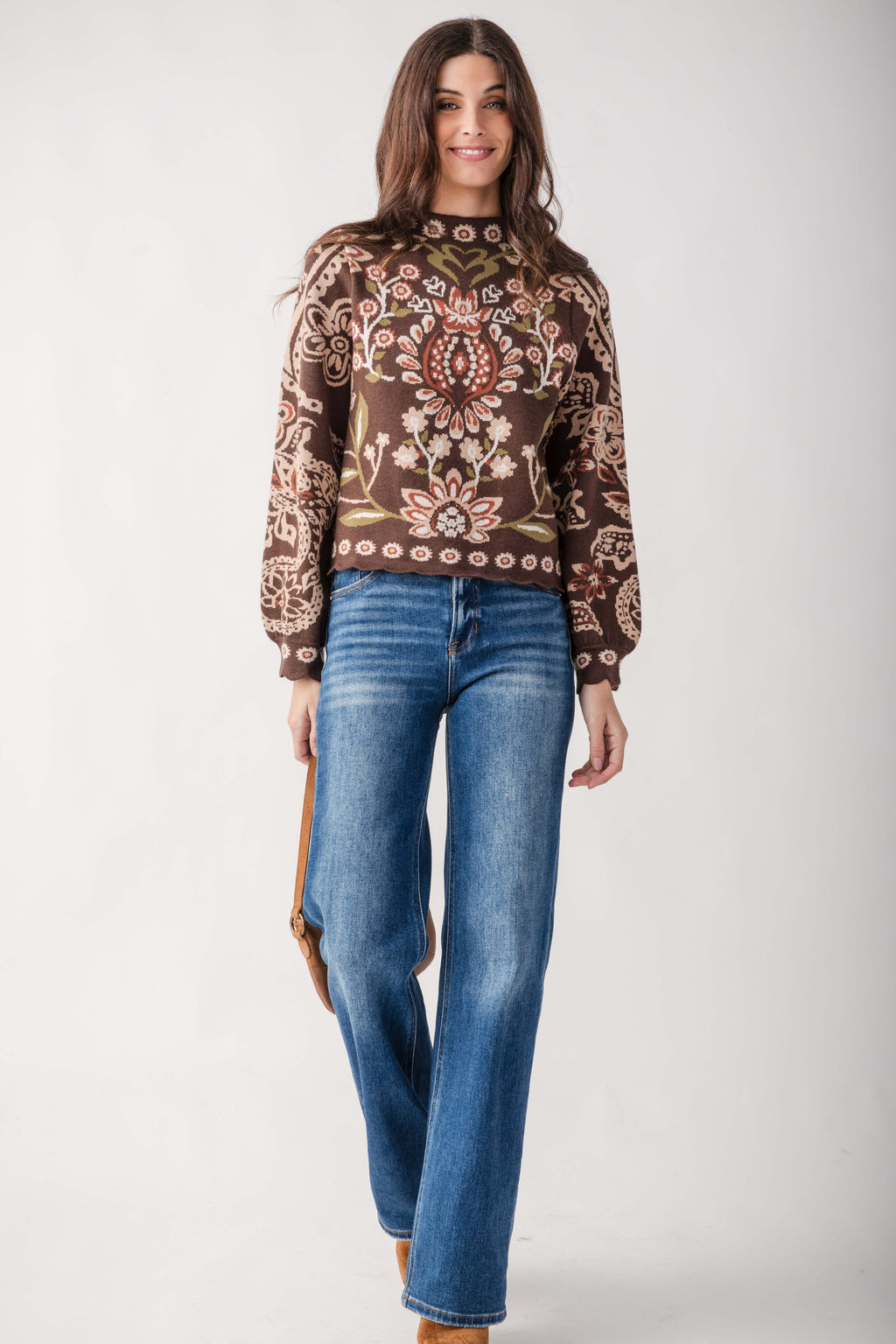A woman modeling a brown long-sleeve knit sweater with an intricate multicolor floral and paisley pattern, paired with blue jeans, against a plain light gray studio background.