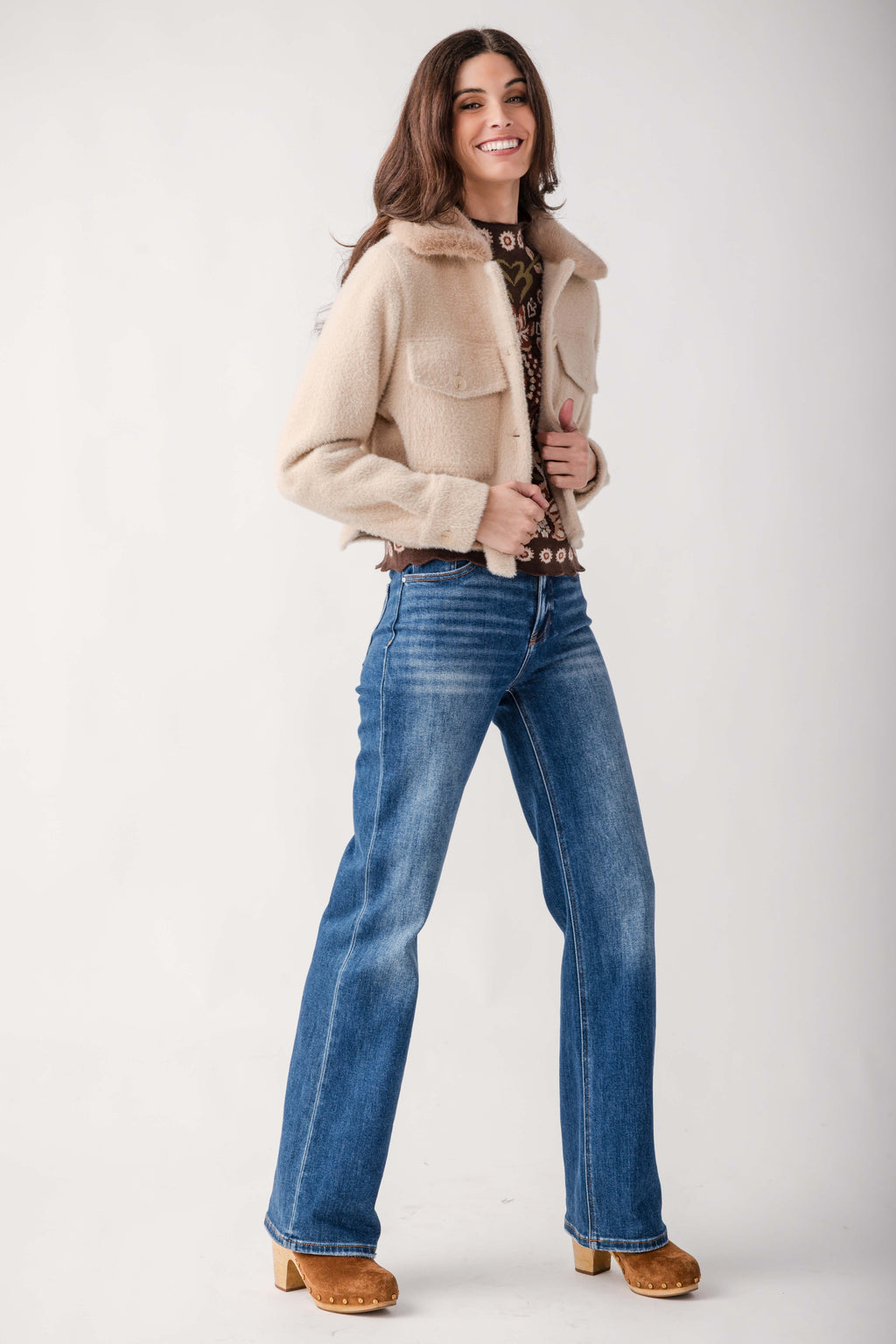 A woman modeling a beige shearling-style cropped jacket, blue straight-leg jeans, a brown patterned top, and brown suede boots against a light gray studio background.