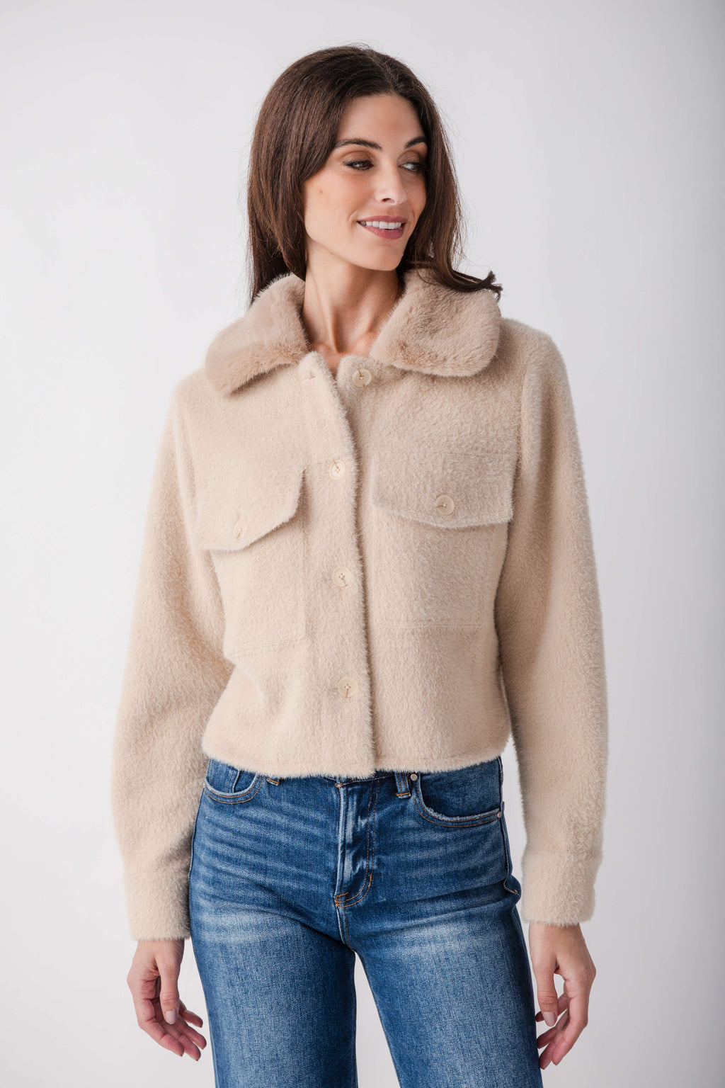 A woman wearing a cropped, fuzzy beige sherpa jacket with a wide collar and button front, paired with blue jeans, against a plain light gray studio background.