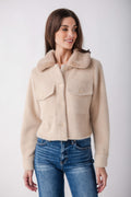 A woman wearing a cropped, fuzzy beige sherpa jacket with a wide collar and button front, paired with blue jeans, against a plain light gray studio background.