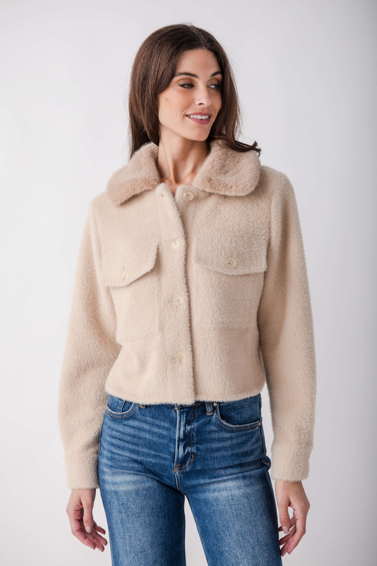 A woman wearing a cropped, fuzzy beige sherpa jacket with a wide collar and button front, paired with blue jeans, against a plain light gray studio background.