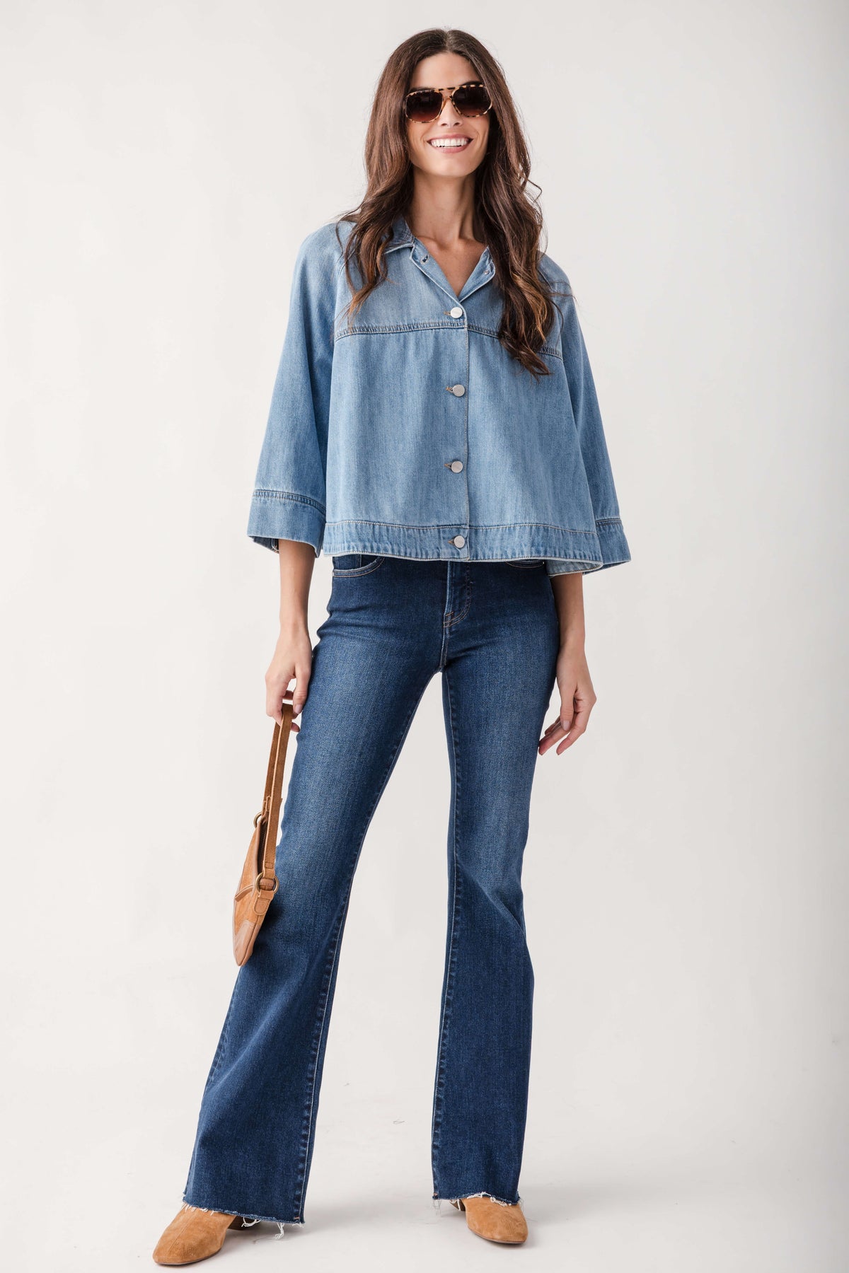 A woman modeling a light-wash blue cropped denim jacket with three-quarter sleeves, paired with dark-wash flare jeans, tan suede shoes, and holding a tan shoulder bag against a light gray studio background.