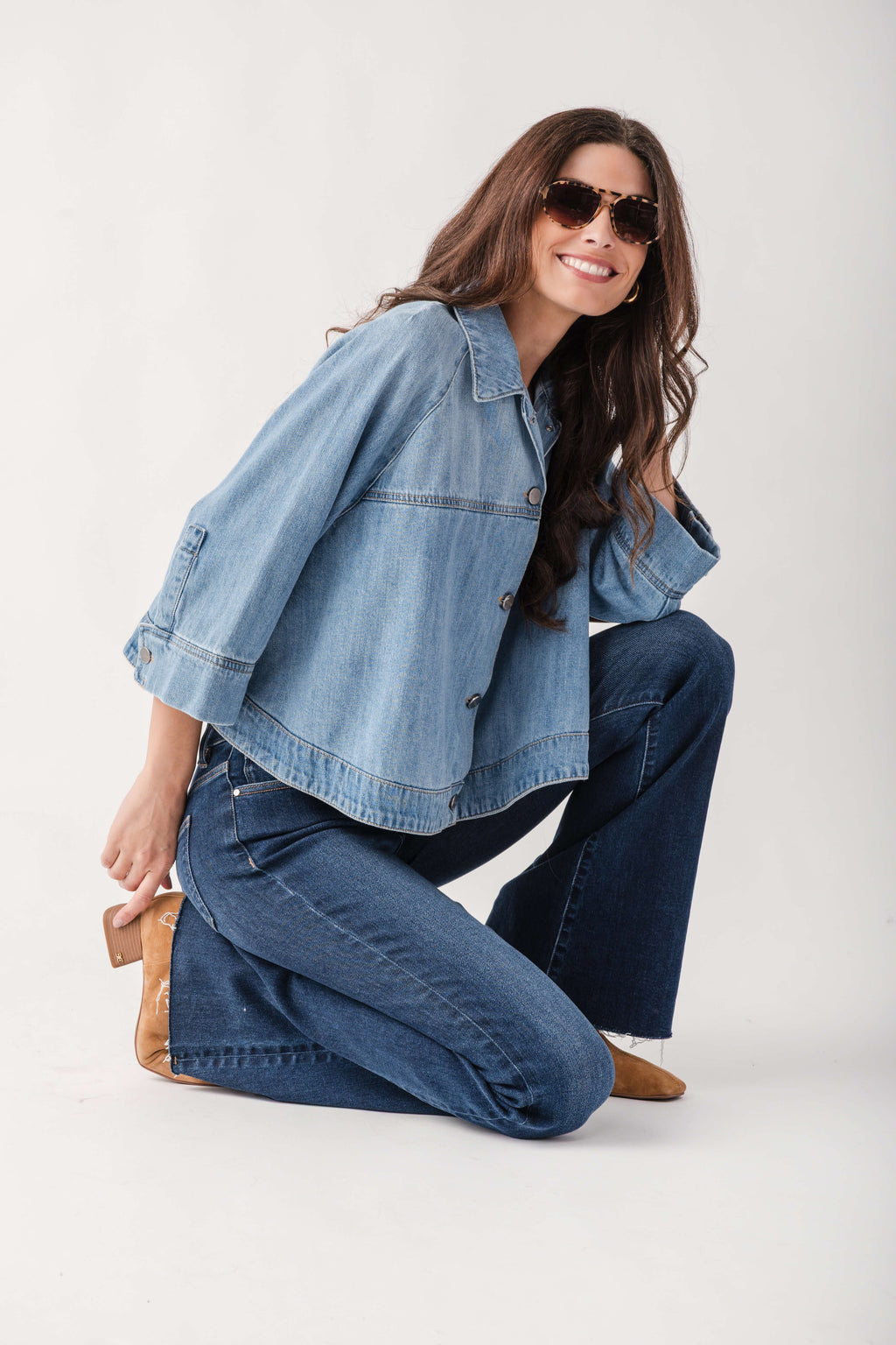 A woman wearing a light blue cropped denim jacket and dark blue wide-leg jeans kneels on a white studio background. She is wearing brown block-heel mules and dark sunglasses.