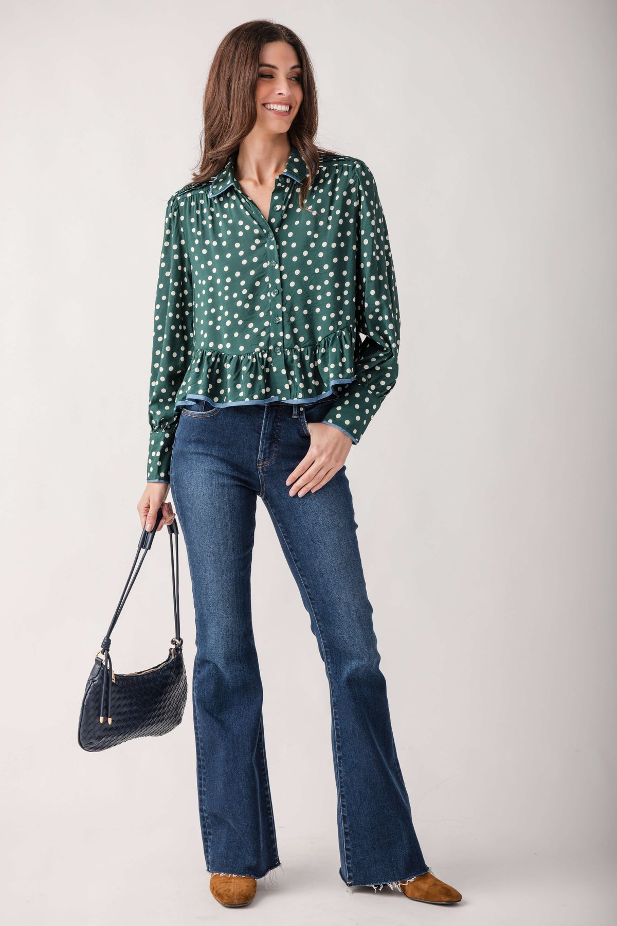 A woman modeling a green long-sleeve blouse with white polka dots, paired with blue flared jeans, brown shoes, and a dark blue handbag, against a light gray studio background.
