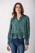 A woman modeling a green long-sleeve blouse with a white polka dot pattern and a ruffled hem, paired with blue jeans, against a light gray studio background.