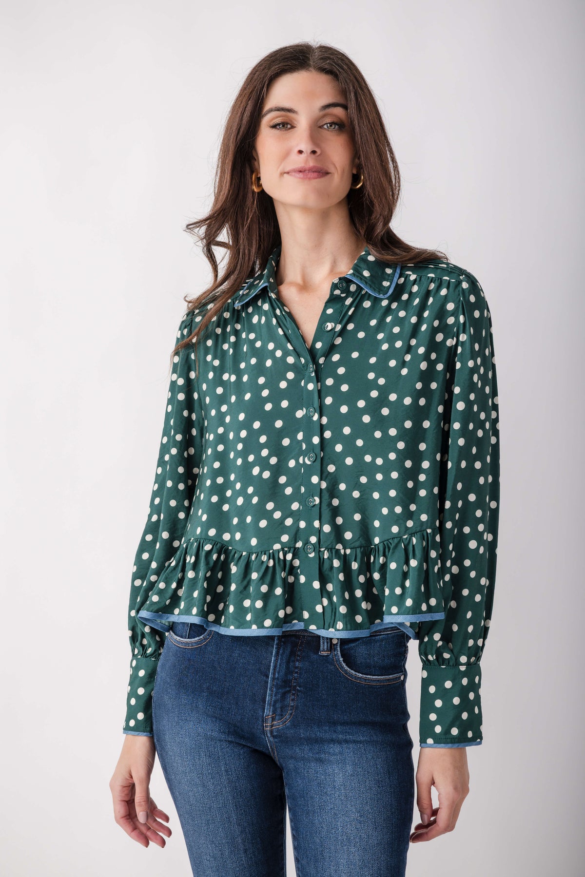 A woman modeling a green long-sleeve blouse with a white polka dot pattern and a ruffled hem, paired with blue jeans, against a light gray studio background.