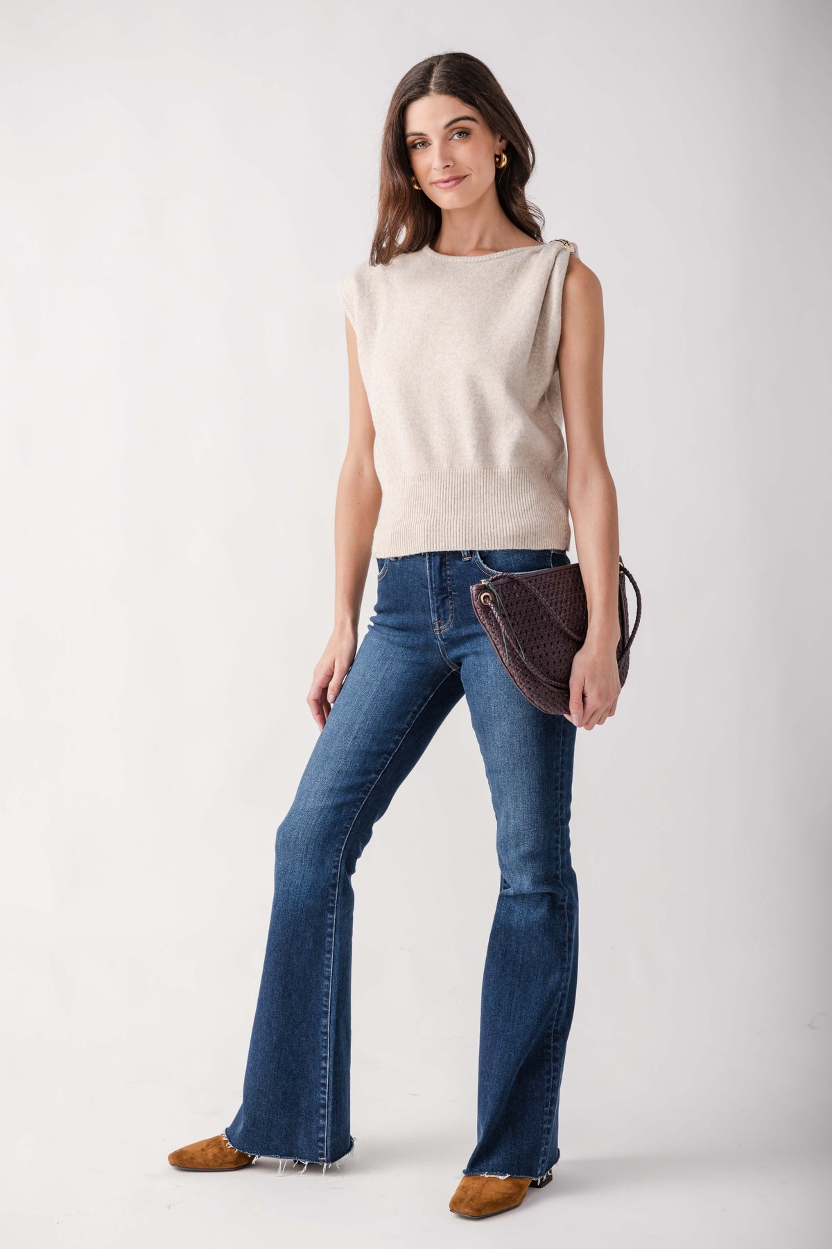 A woman modeling a sleeveless cream knit top, blue flare jeans, and brown suede shoes, holding a dark brown textured shoulder bag against a plain light gray studio background.