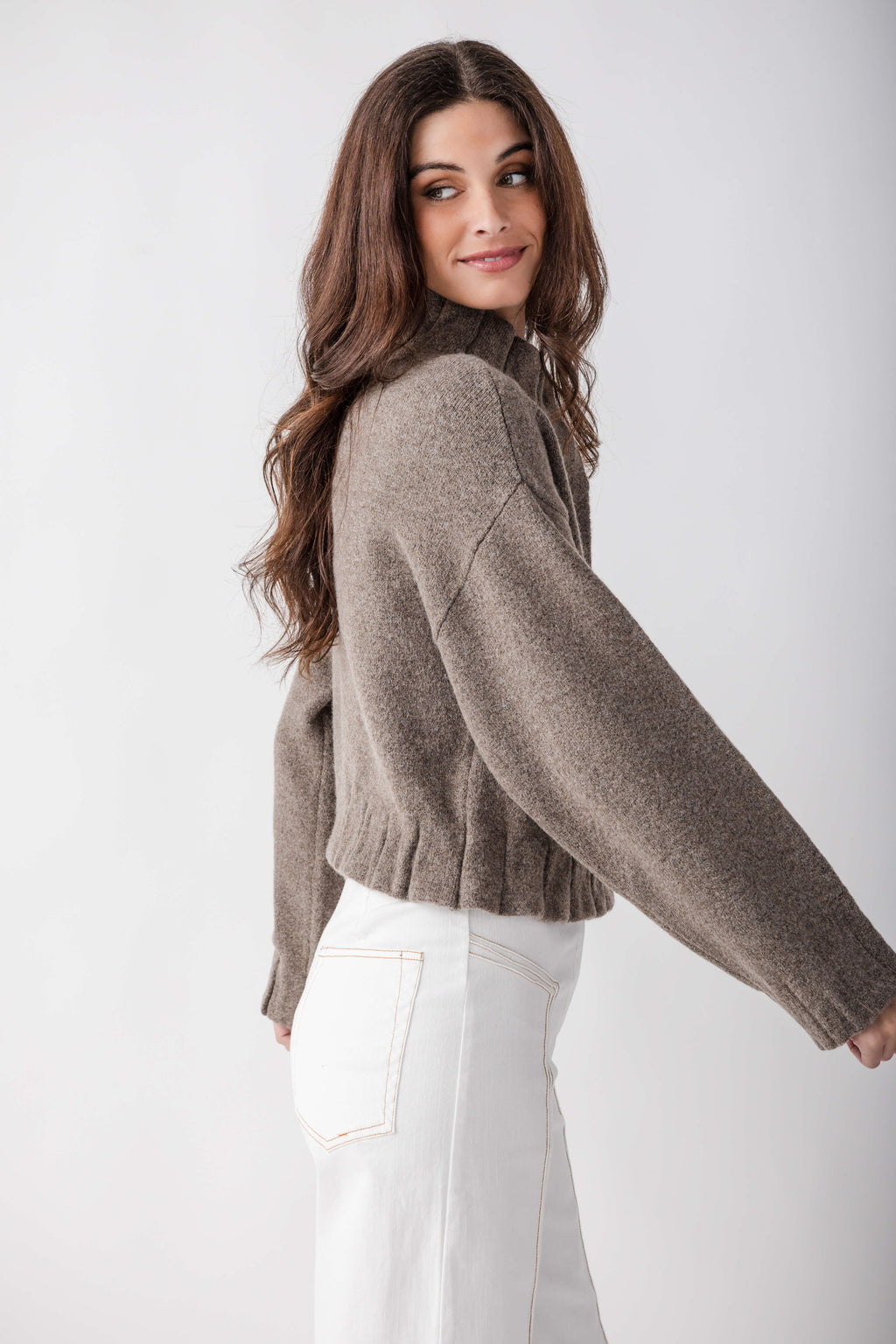 Taupe knit jacket with pleated ruffle hem worn over white jeans, shown from back angle.