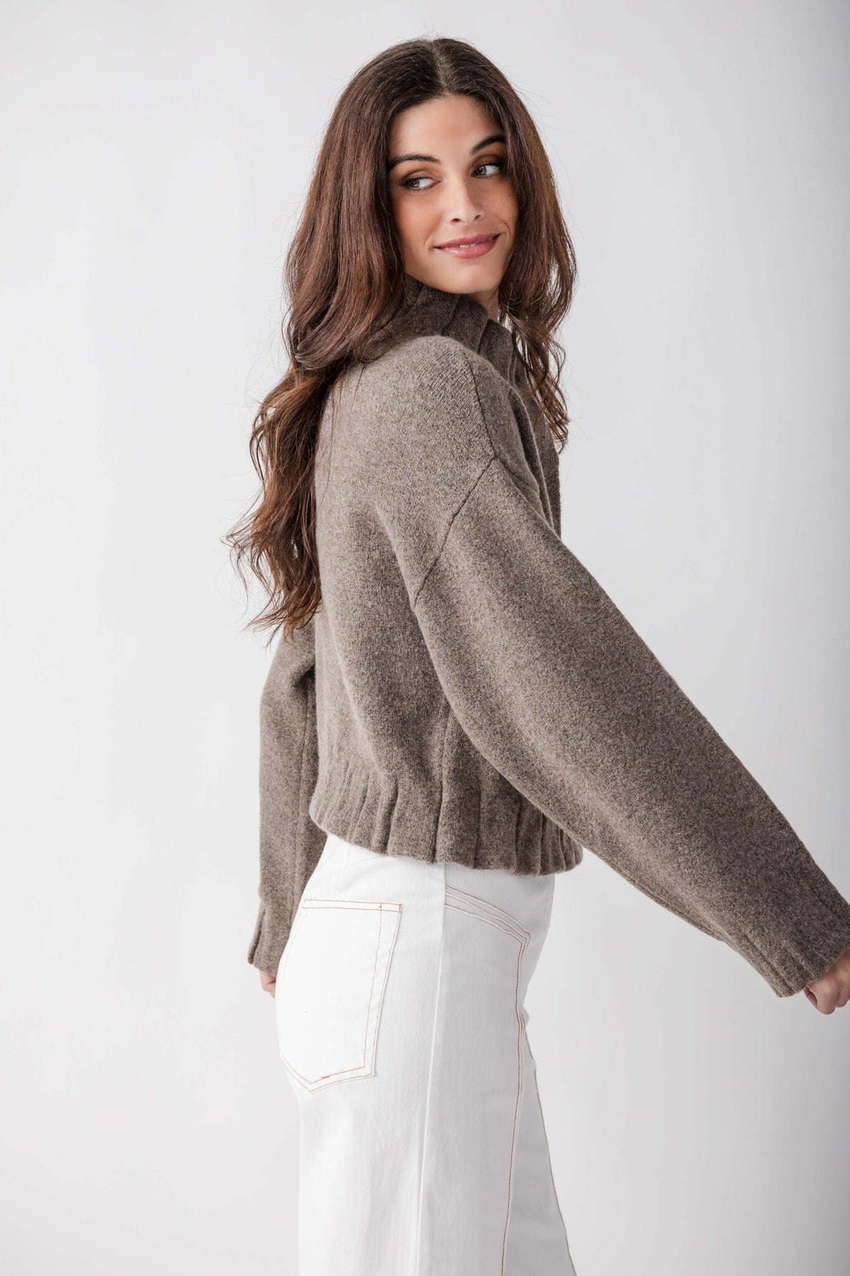 Taupe knit jacket with pleated ruffle hem worn over white jeans, shown from back angle.